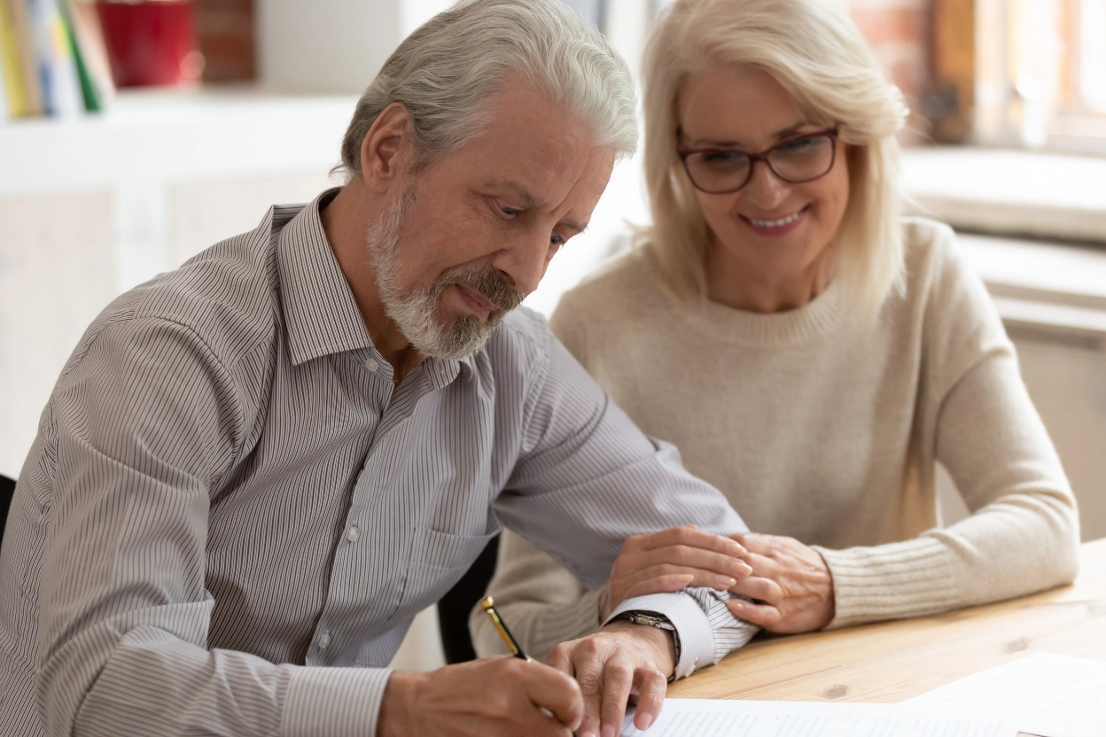 Older couple reviewing and signing estate planning documents together at a table, with the woman gently supporting the man as he writes.