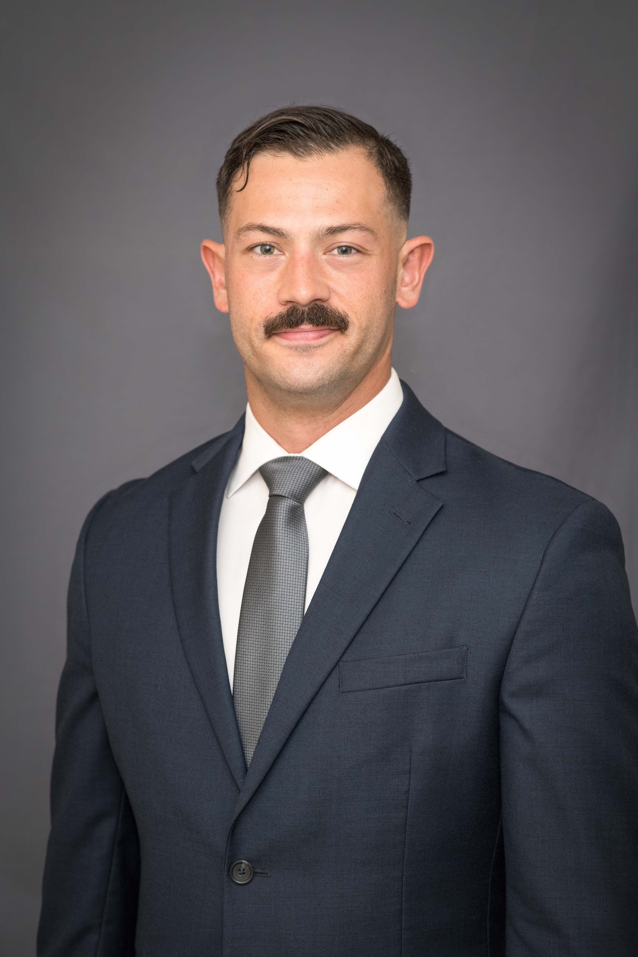 Portrait of a man with a mustache wearing a dark suit, white shirt, and gray tie against a gray background.