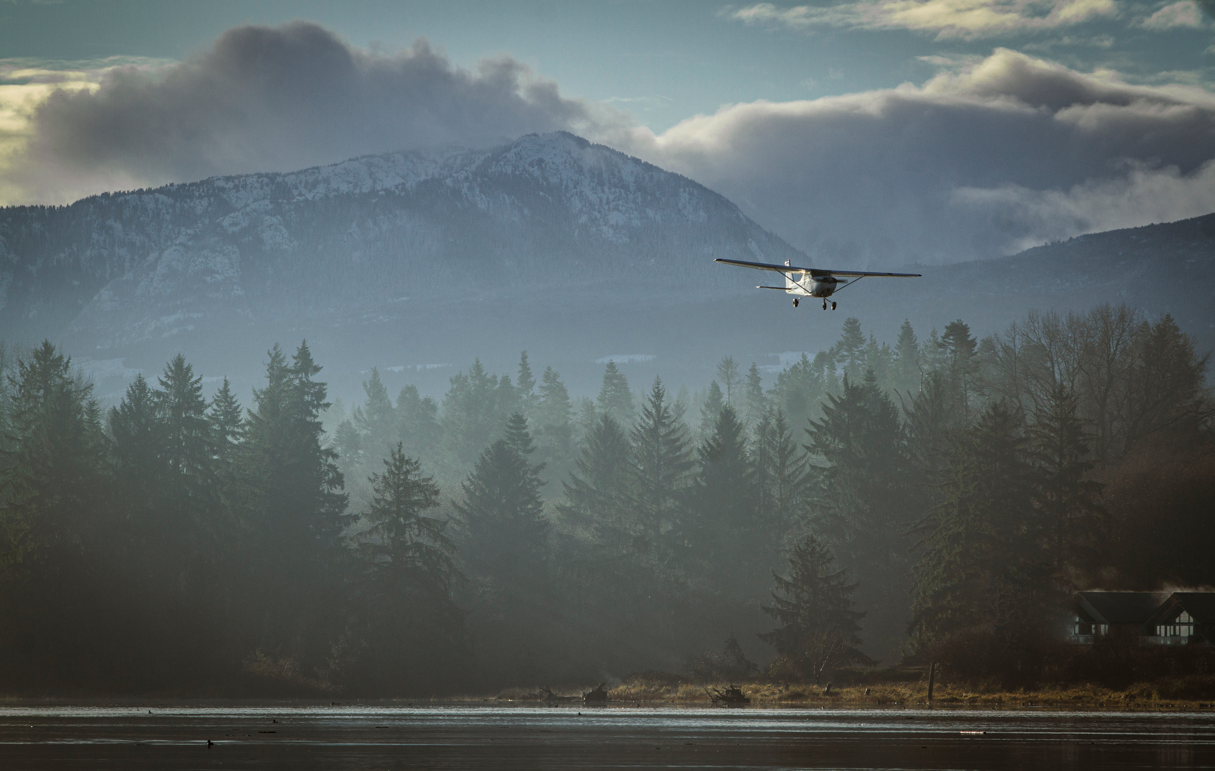 Small airplane flying over misty forest with mountains and cloudy sky in the background.
