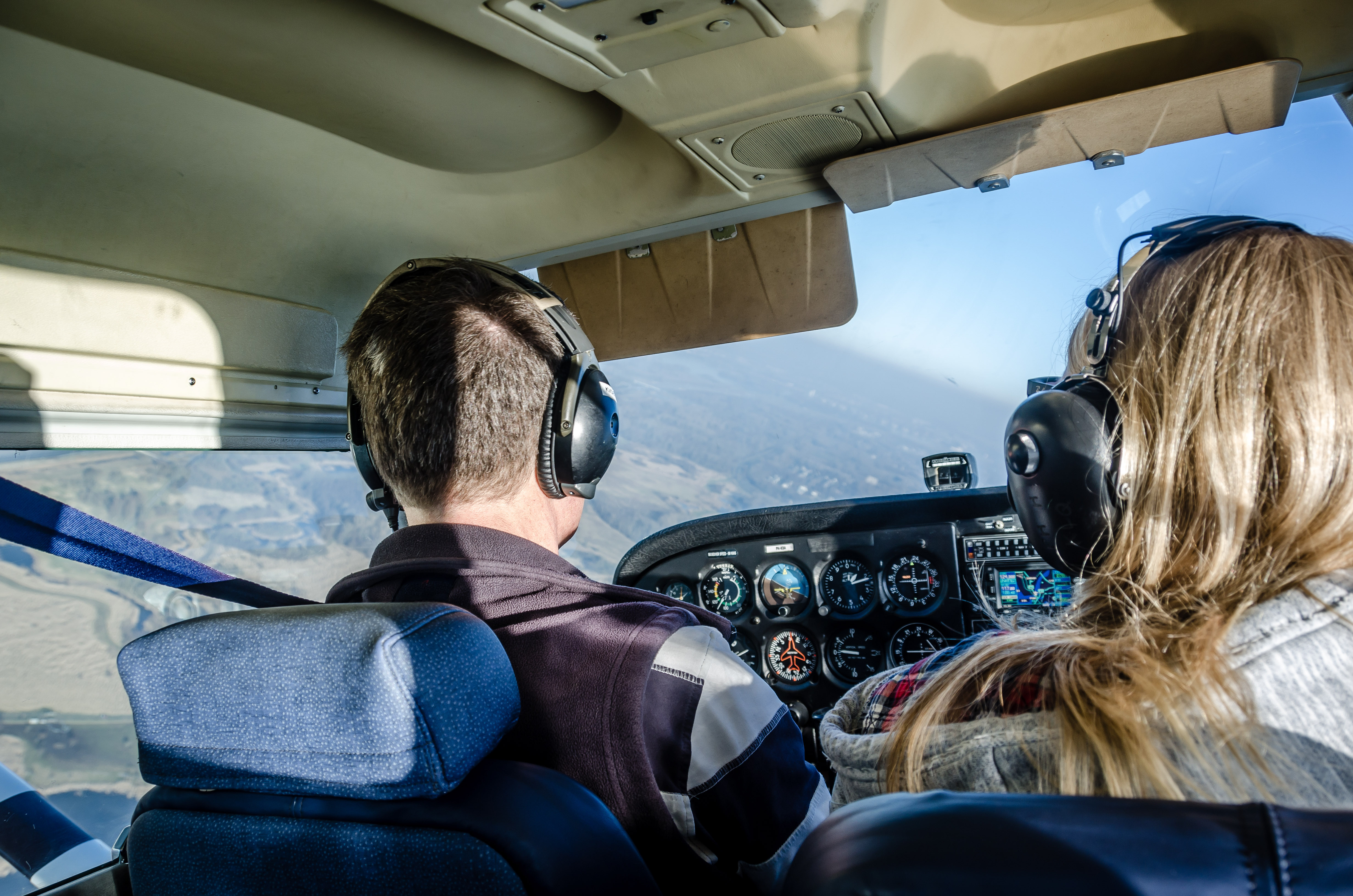 Two people wearing headsets sitting in the cockpit of a small airplane flying over a mountainous landscape.