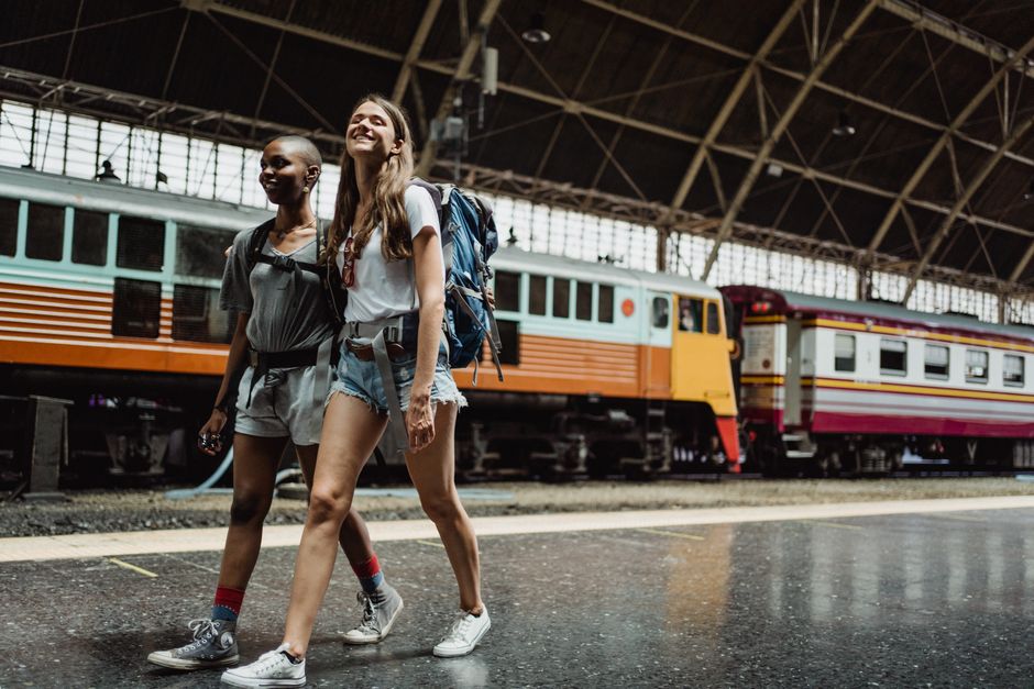 Two women with backpacks walking and smiling at a railway station.