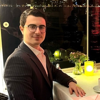 Man with glasses in a dark pinstripe suit sitting at a restaurant table with a vase of flowers and candle, city lights reflected on the window behind him.