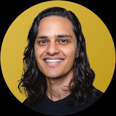 Smiling man with long black hair wearing a black shirt against a golden yellow circular background.