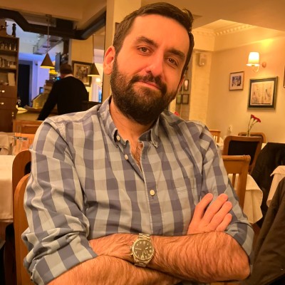Man with a beard and short hair wearing a blue checkered shirt sitting in a restaurant with arms crossed and a slight smile.
