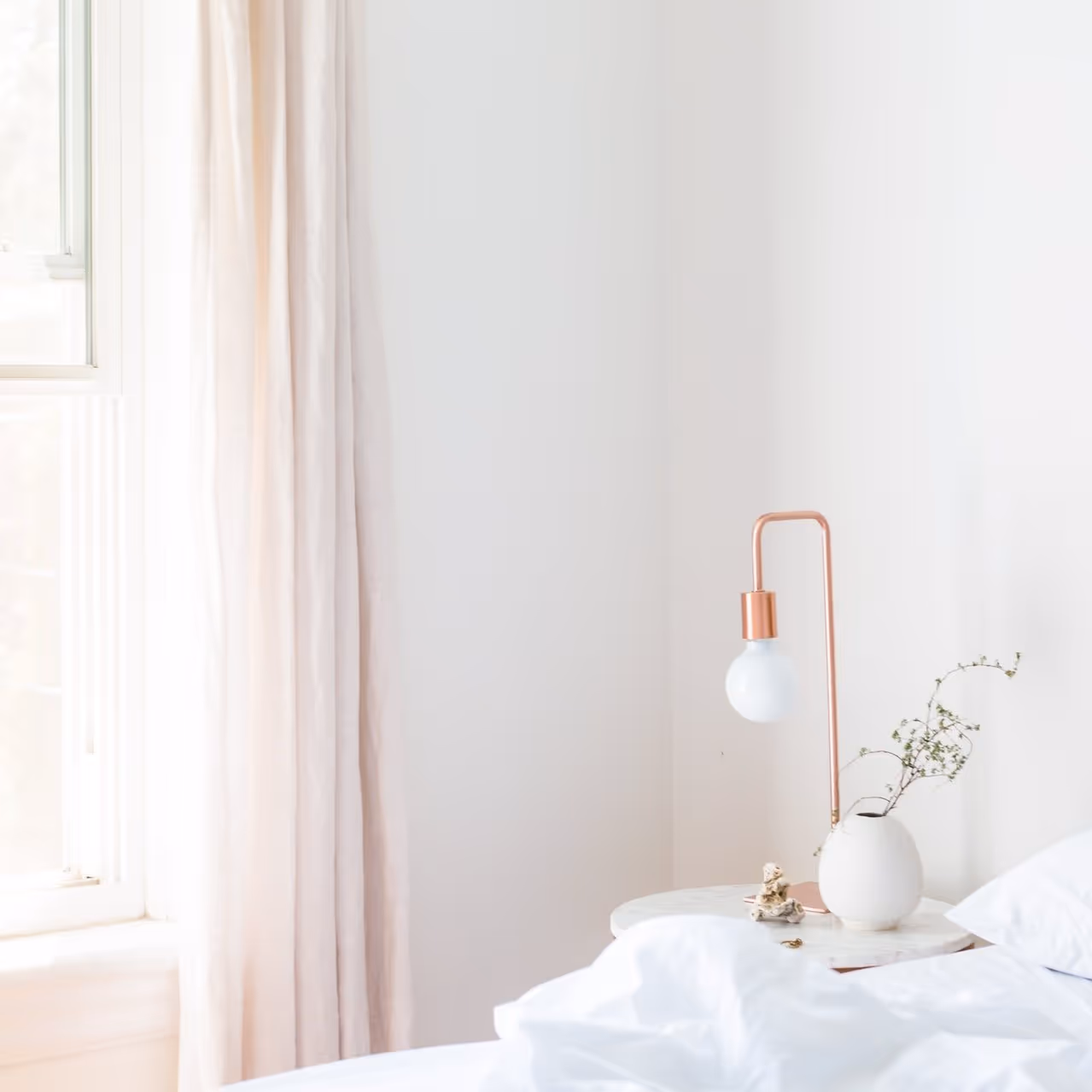 Minimalistisk soveromshjørne med hvitt sengetøy, en kobberlampe og en vase på nattbordet.