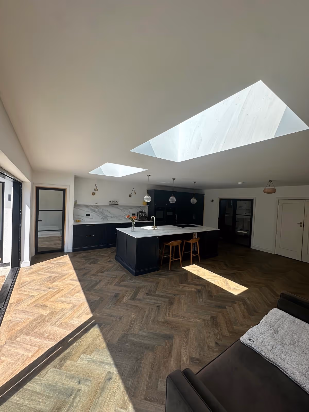 Modern kitchen with black cabinets, marble countertops, three hanging pendant lights, two skylights, and herringbone wood flooring.