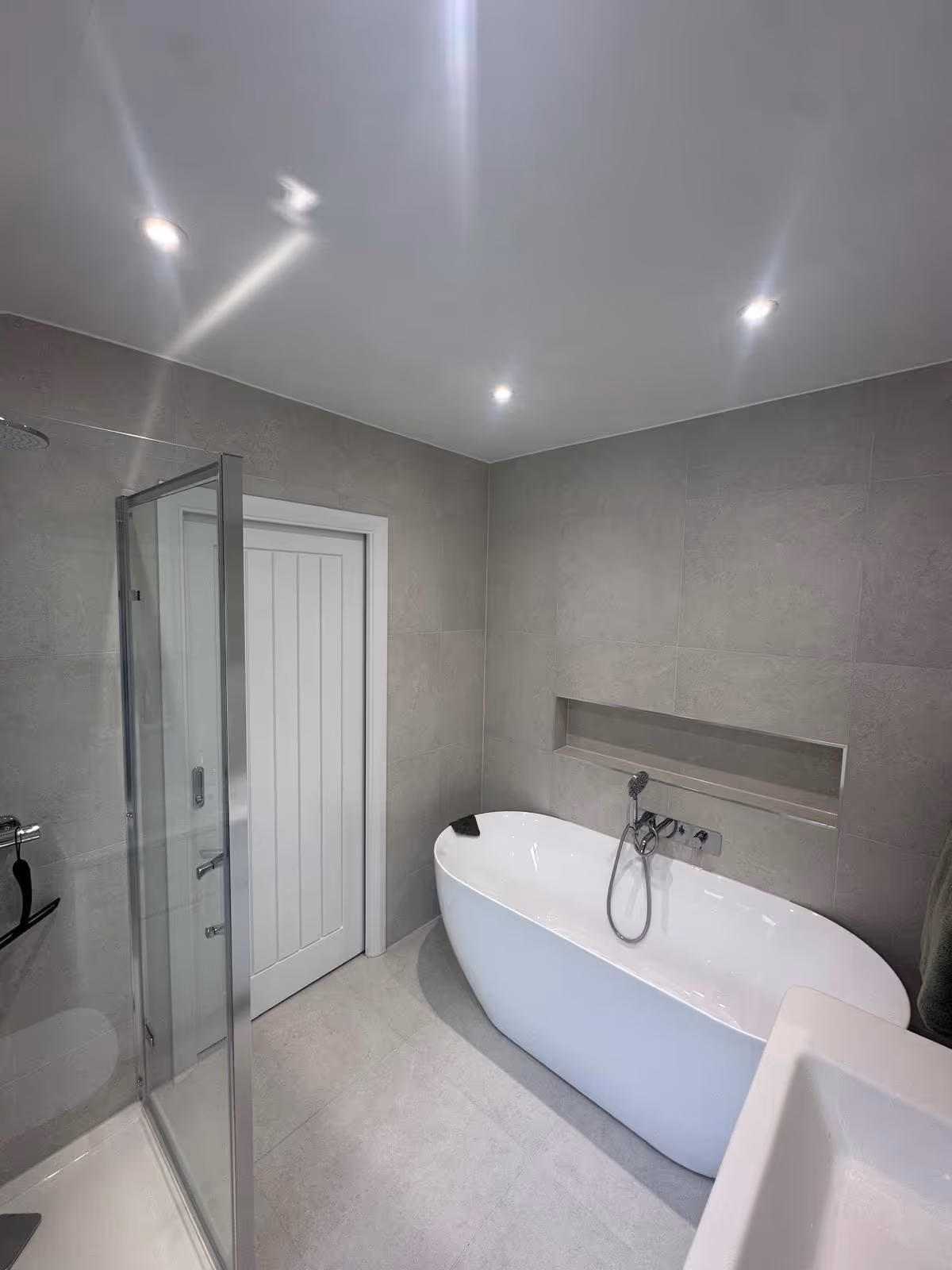 Modern bathroom with a freestanding white bathtub, glass-enclosed shower, white door, and gray tiled walls and floor.