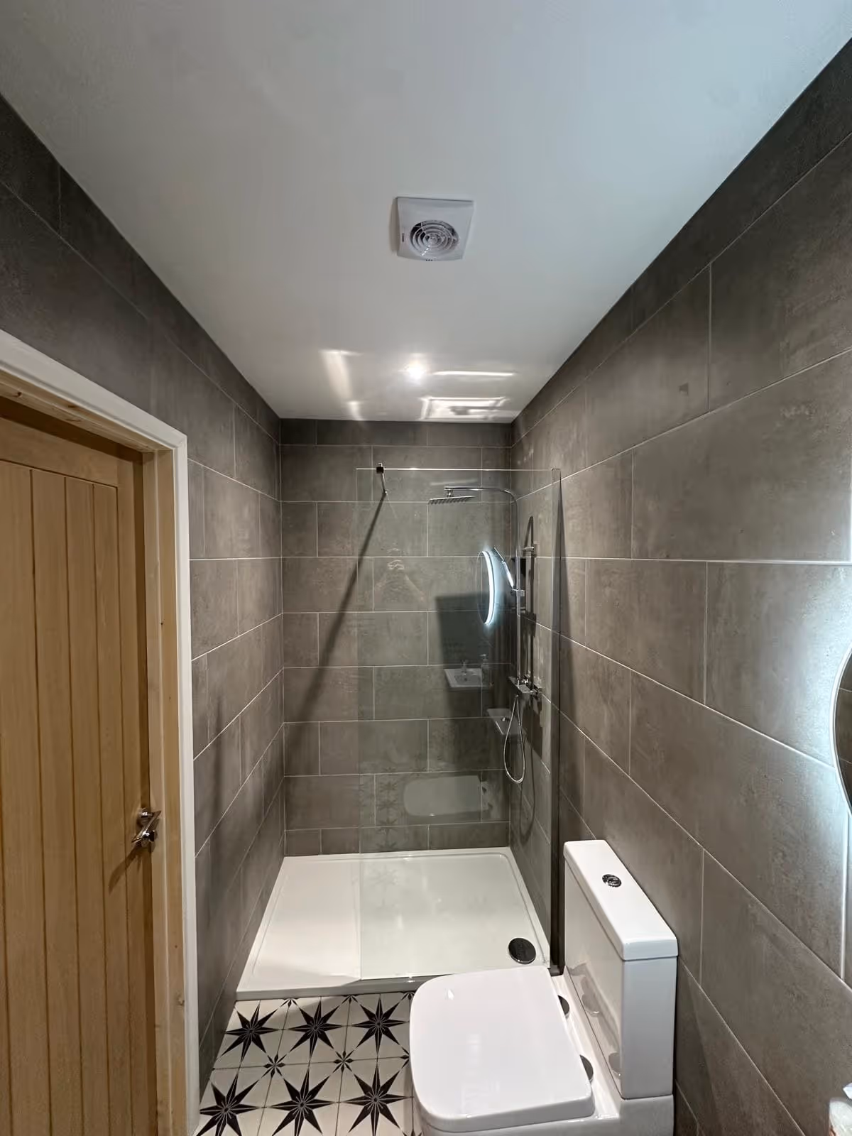 Modern bathroom with gray tiled walls, patterned floor tiles, walk-in glass shower, and white toilet.