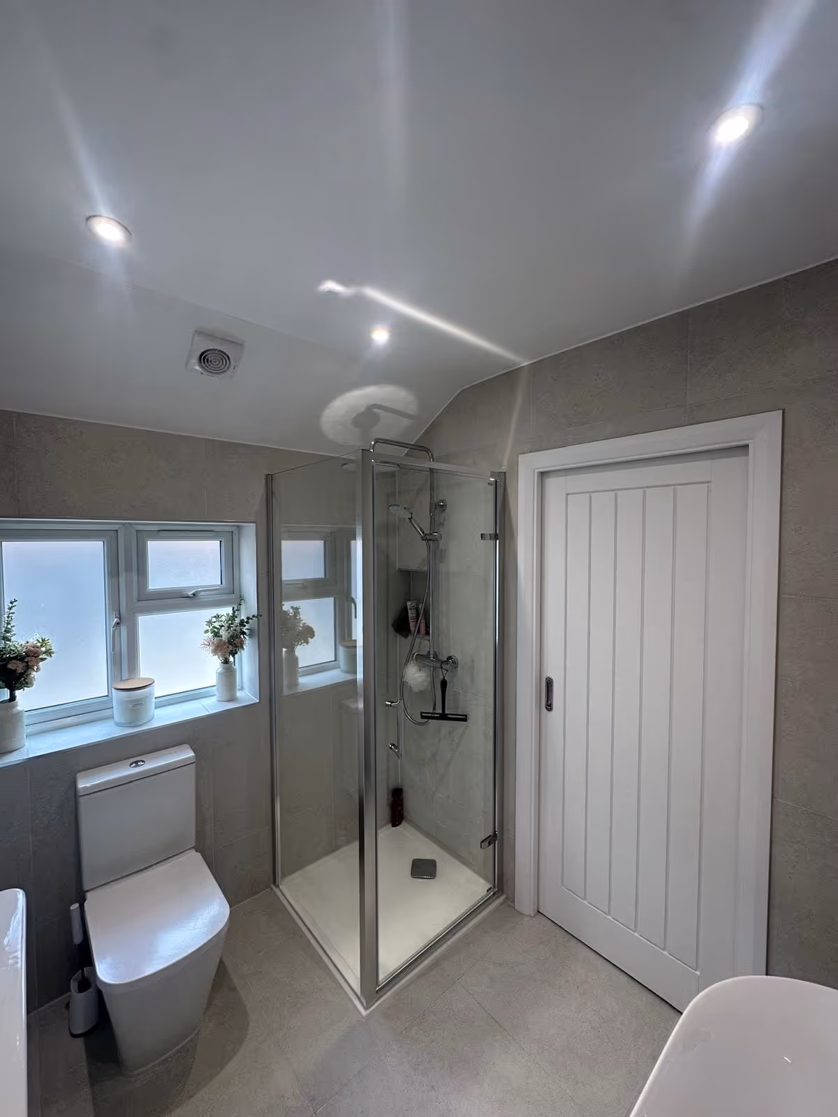 Modern bathroom with glass shower enclosure, white toilet, frosted window with flower pots, and a white sliding door.