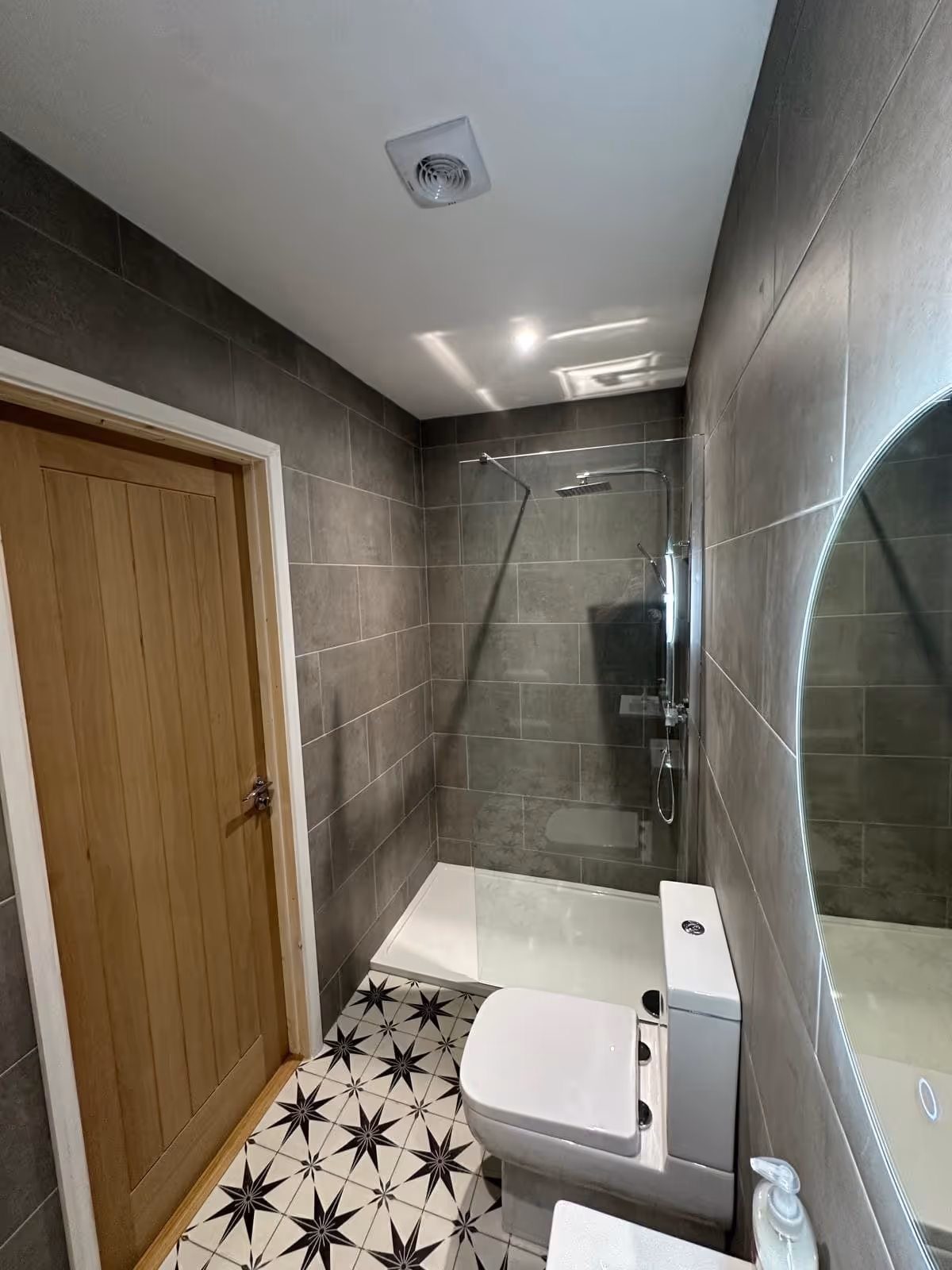 Modern bathroom with gray tiled walls, patterned black and white floor tiles, a glass-enclosed shower, white toilet, wooden door, and round illuminated mirror.