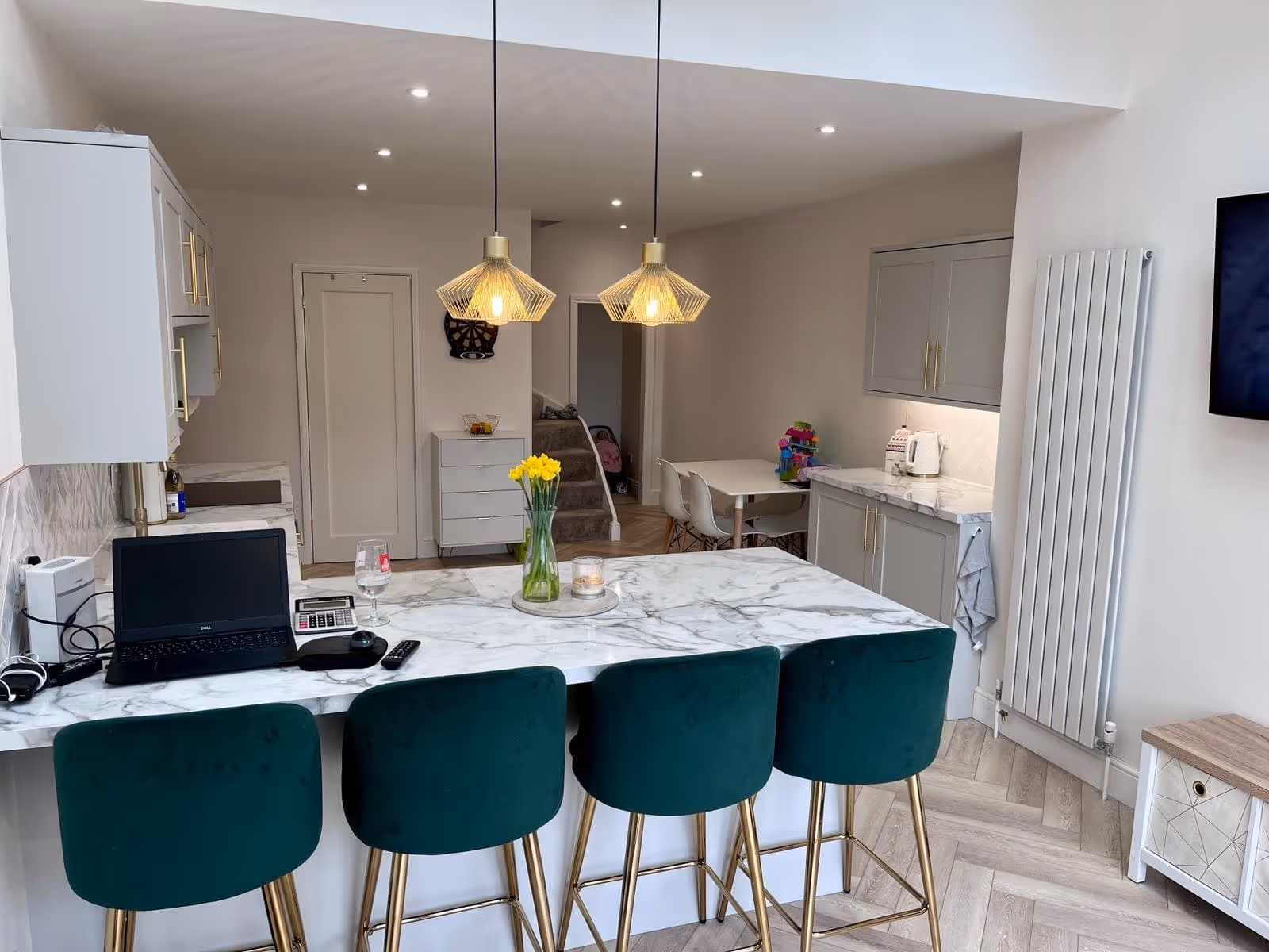 Modern kitchen with marble countertops, four green bar stools, pendant lights, a laptop, and a vase of yellow flowers on the island.