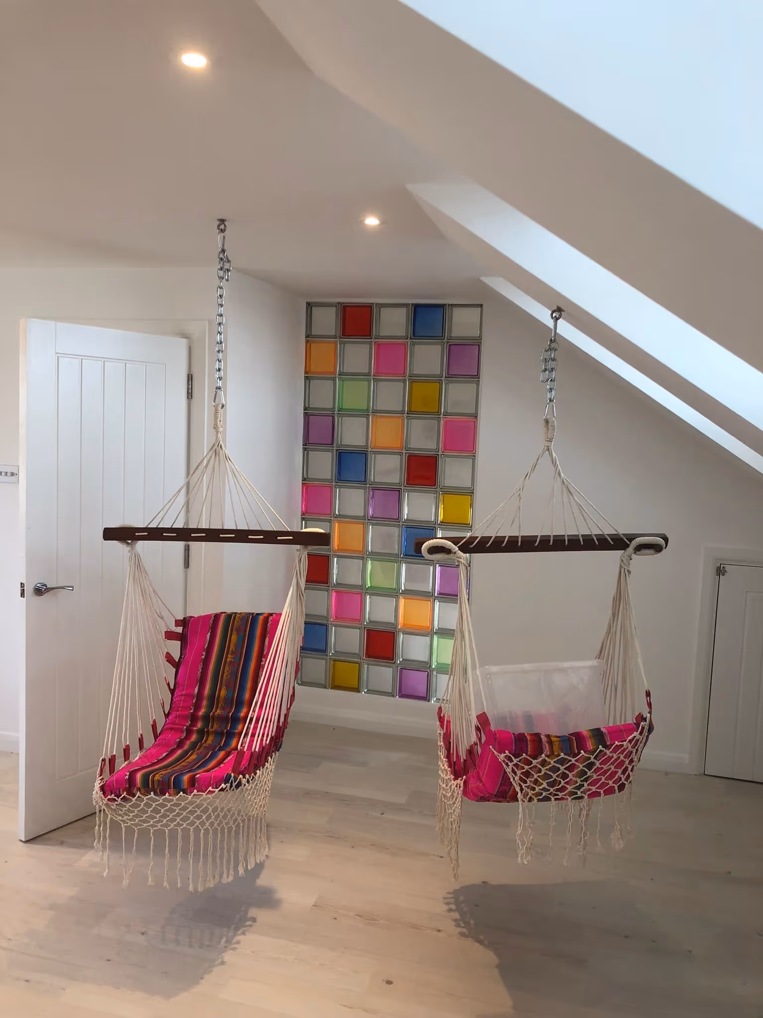 Two hanging hammock chairs with colorful striped cushions in a bright room with slanted ceiling and multicolored glass block wall.
