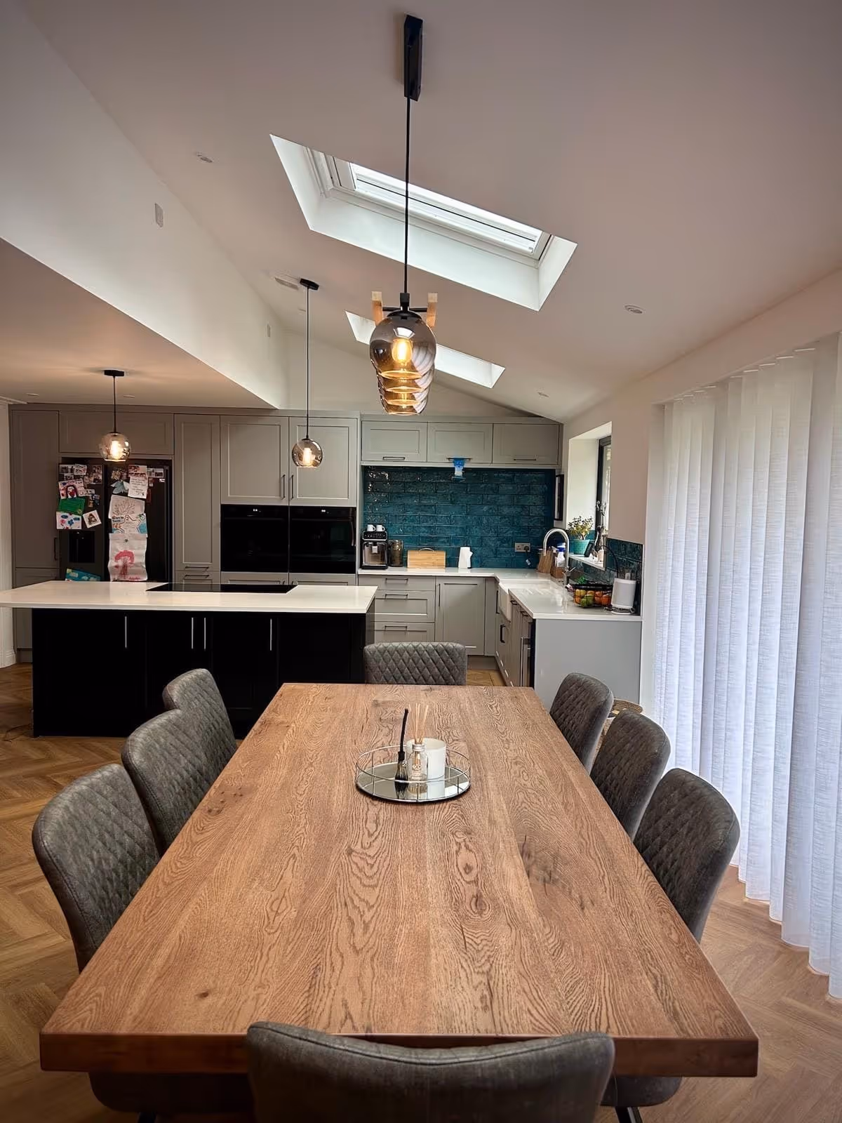 Modern kitchen with wooden dining table, six gray upholstered chairs, pendant lights, skylights, gray cabinets, and teal tile backsplash.
