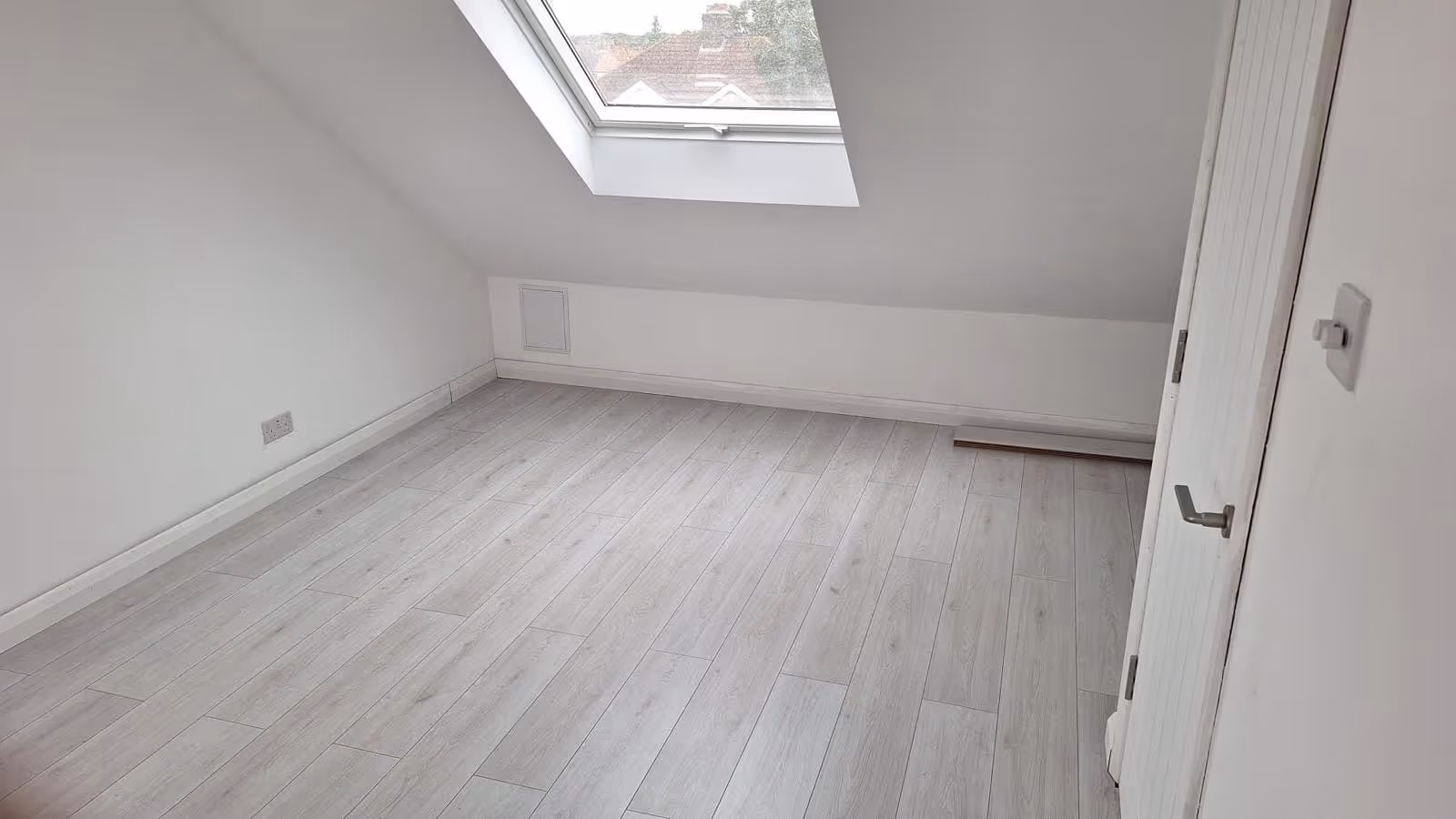 Empty room with light gray wooden flooring, white walls, a skylight window, and a partially open white door.