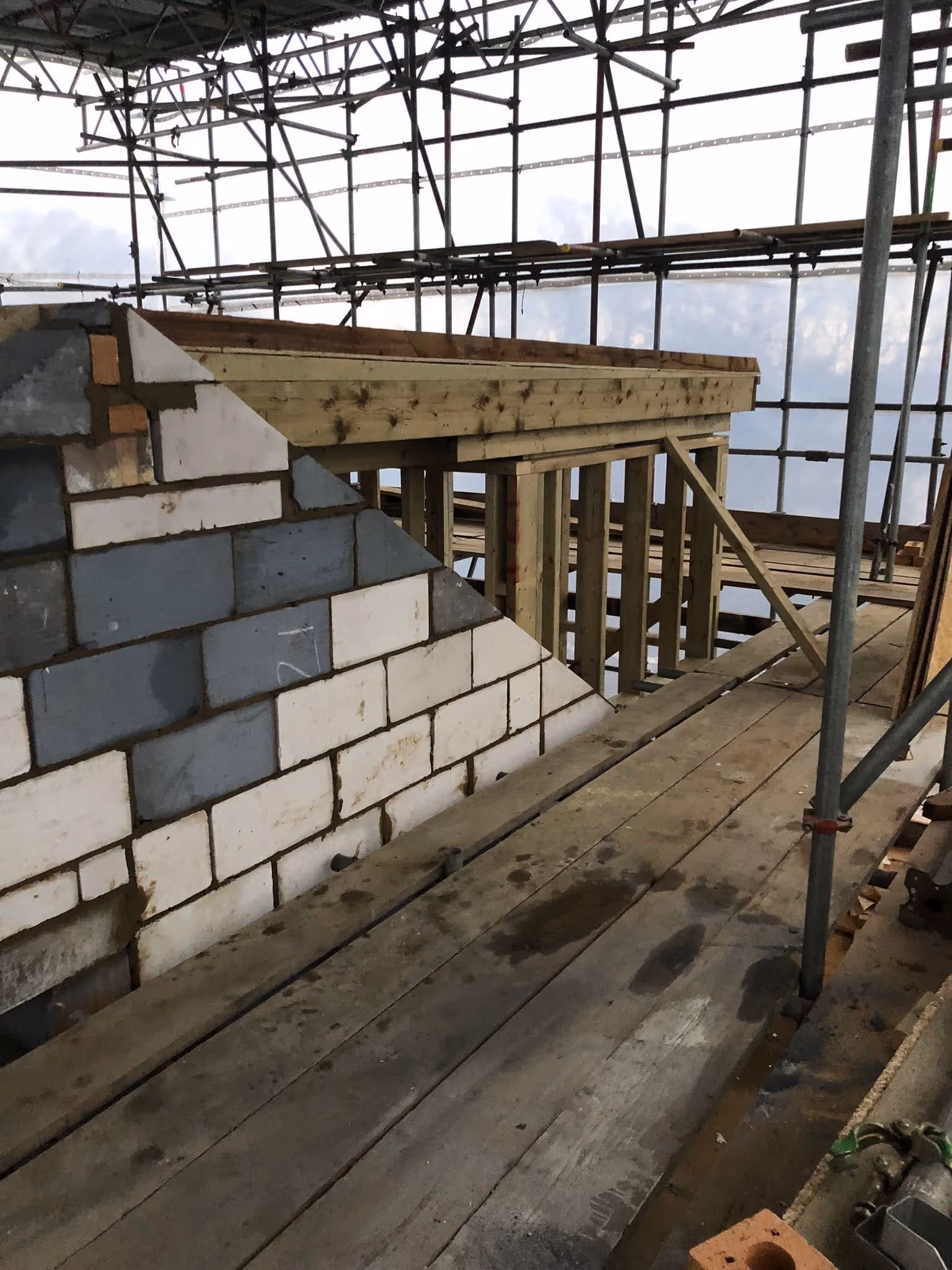 Construction site showing wooden scaffolding and partially built brick wall with concrete blocks.