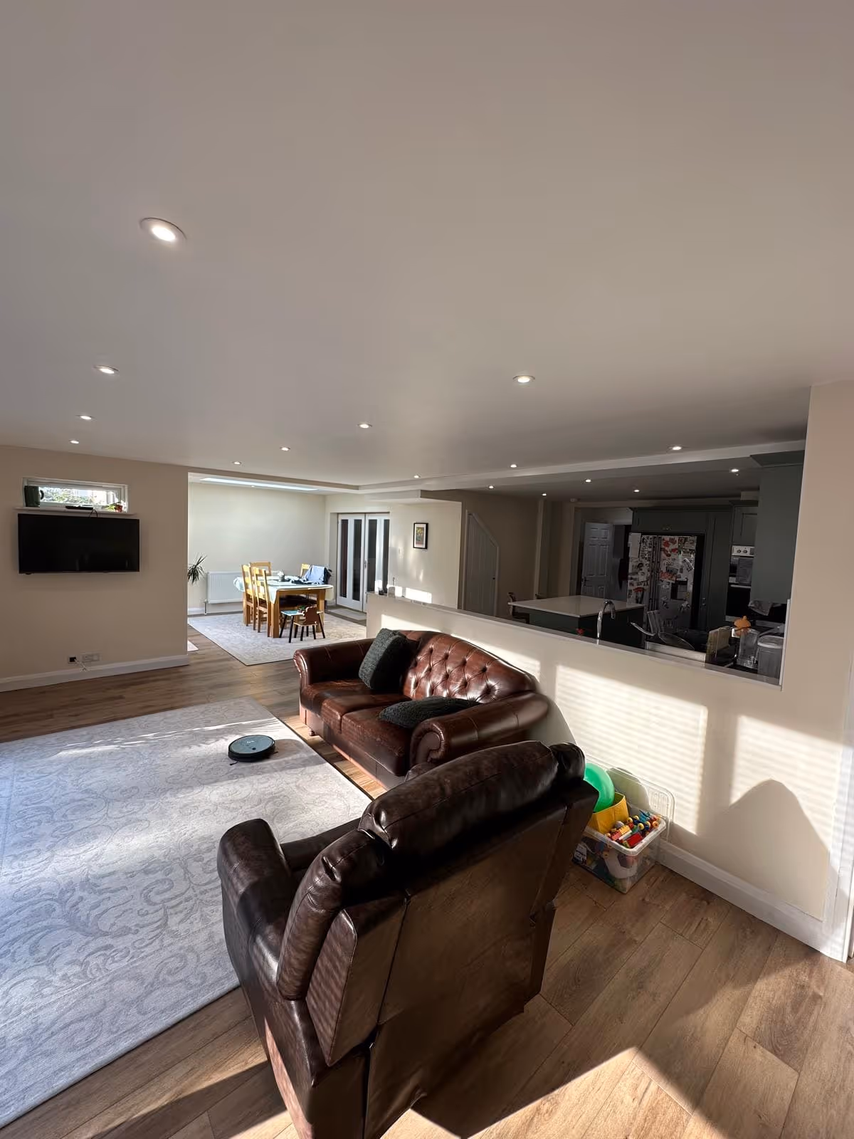 Sunlit living room with two brown leather sofas, a patterned rug, a wall-mounted TV, and a dining area in the background.