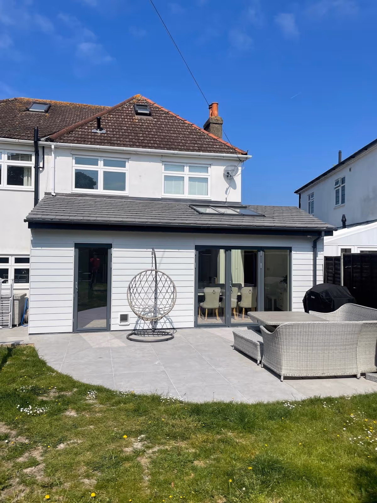 Backyard view of a two-story house with white siding on the extension, patio with gray tiles, hanging egg chair, and wicker outdoor furniture under clear blue sky.