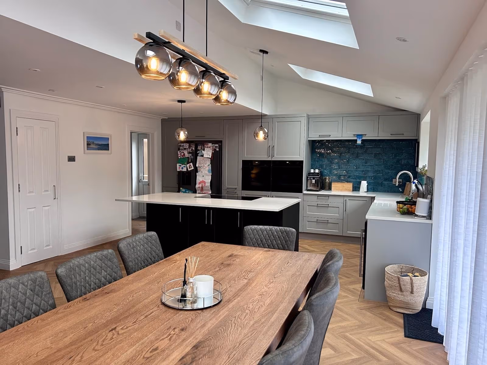 Modern kitchen and dining area with wooden table, gray upholstered chairs, black kitchen island with white countertop, gray cabinets, teal backsplash, and pendant lights.