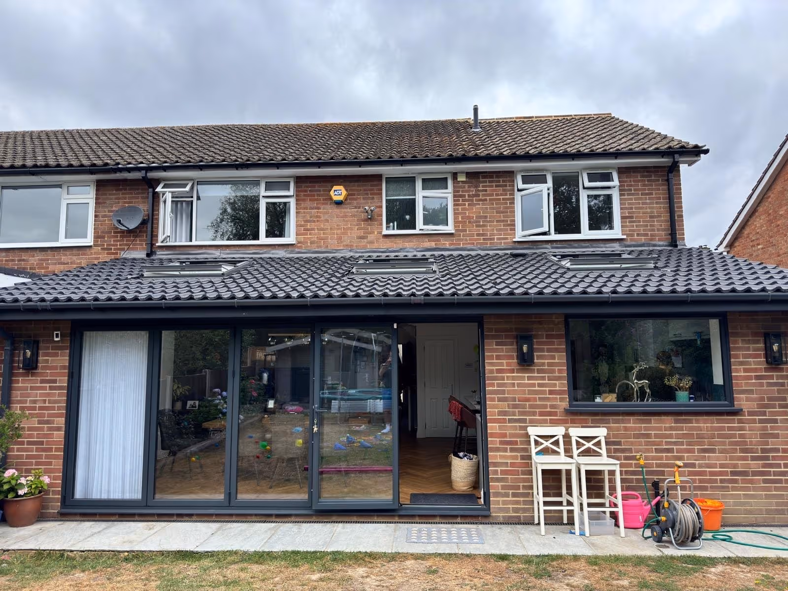 Rear view of a brick two-story house with a tiled roof, large sliding glass doors, two white chairs, and garden hose on a patio.