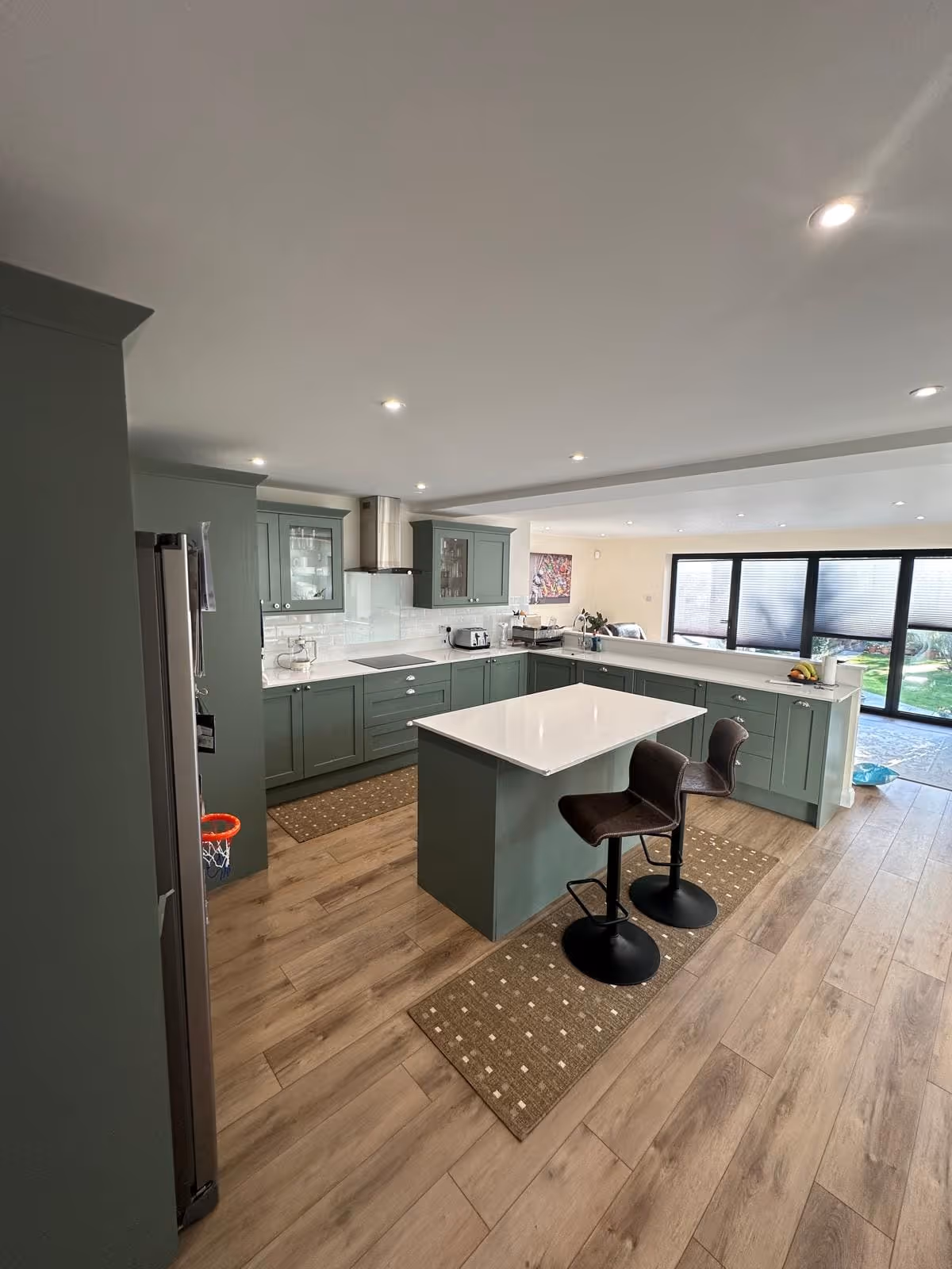 Modern kitchen with green cabinetry, white countertops, kitchen island with two brown bar stools, wooden floor, and large windows overlooking a garden.