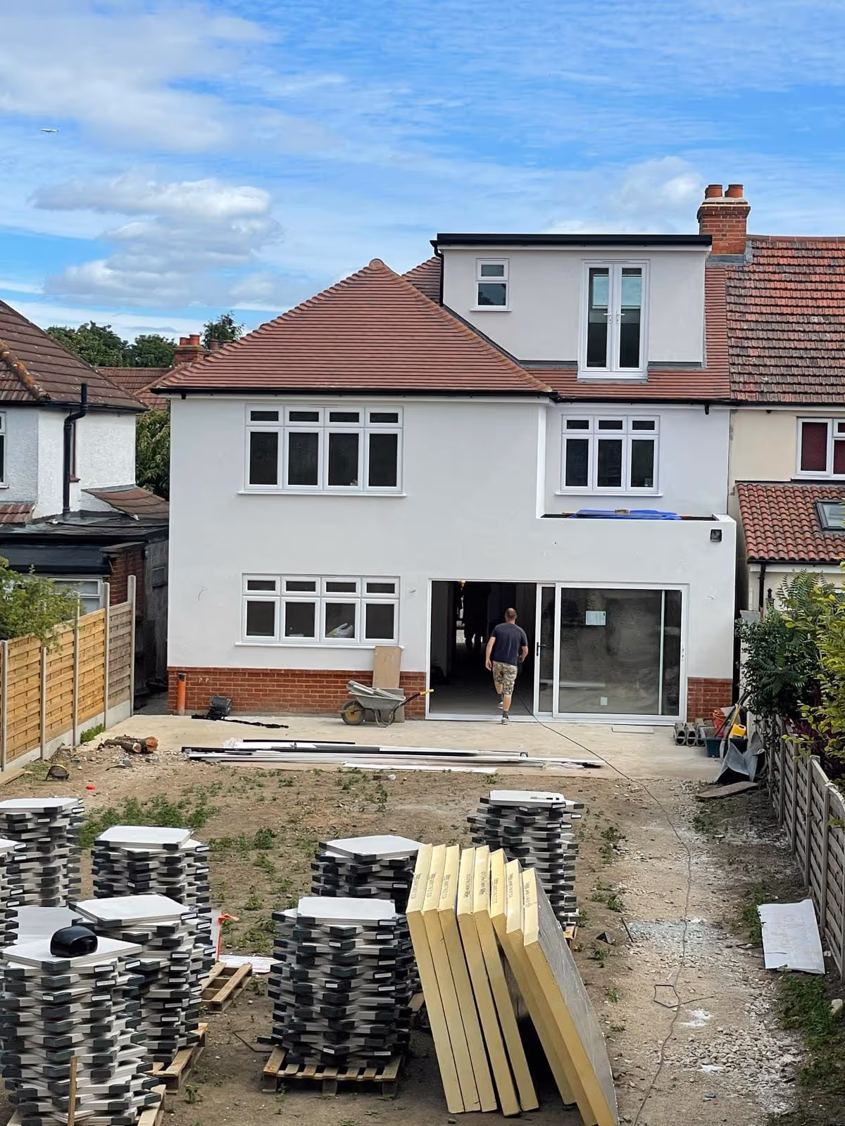Rear view of a two-story house under construction with construction materials stacked on pallets in the backyard and a man walking towards an open sliding door.