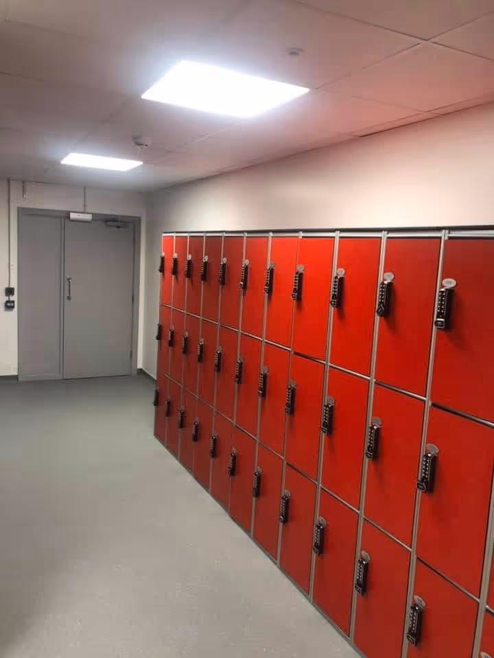 A corridor with a row of red lockers on the right side and a closed double gray door at the end under ceiling lights.