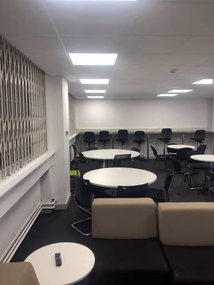 Empty room with multiple round white tables, black chairs, beige sofas, and fluorescent ceiling lights.