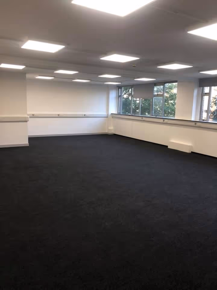 Empty office room with black carpet, white walls, ceiling lights, and windows showing trees outside.