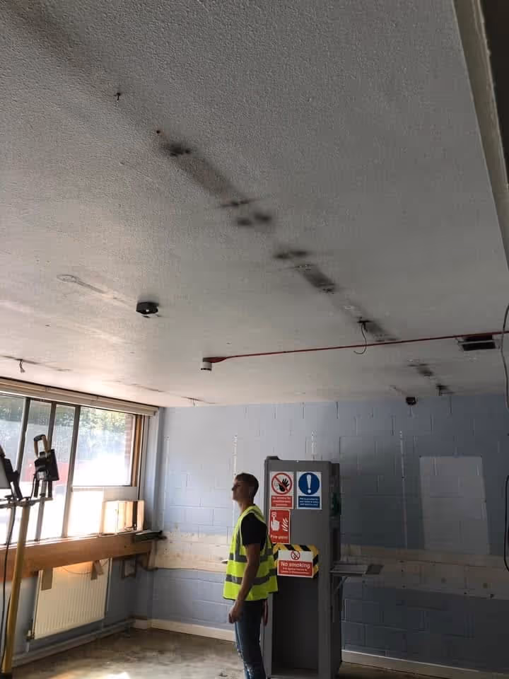 Man in yellow safety vest inspecting a ceiling with dark smoke stains in an industrial room with equipment and safety signs.
