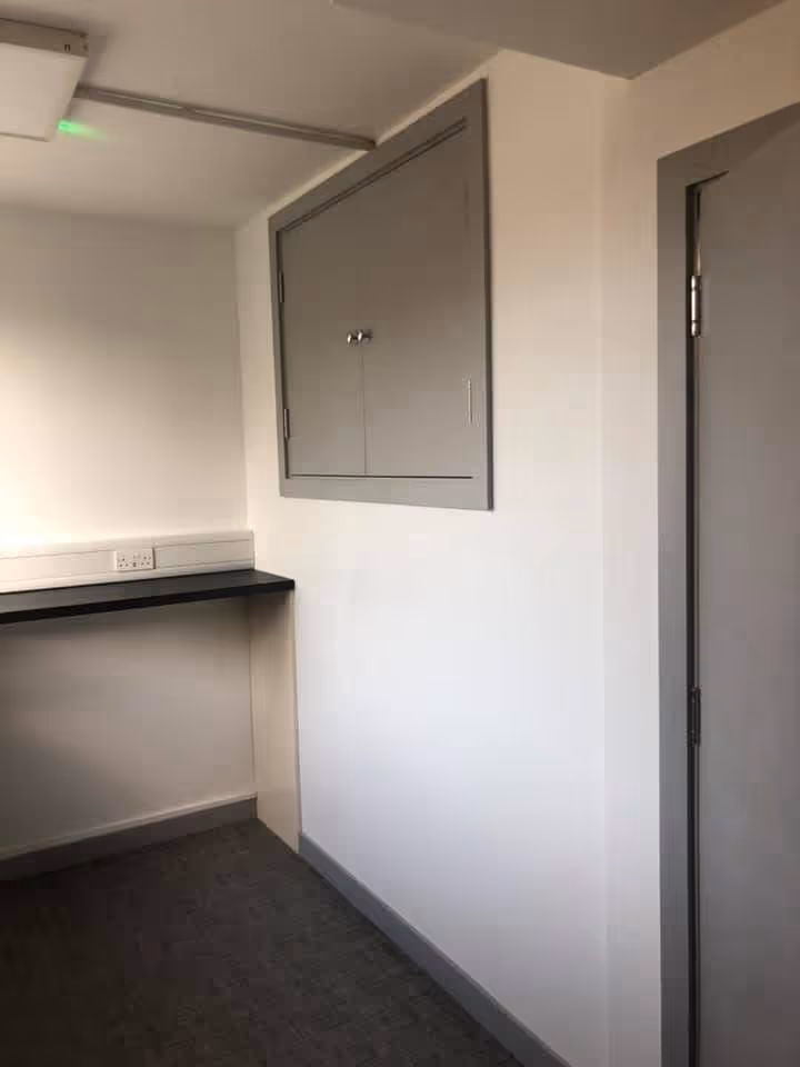 Small corner room with white walls, a gray utility cabinet, a gray door, and a black countertop with electrical outlets.