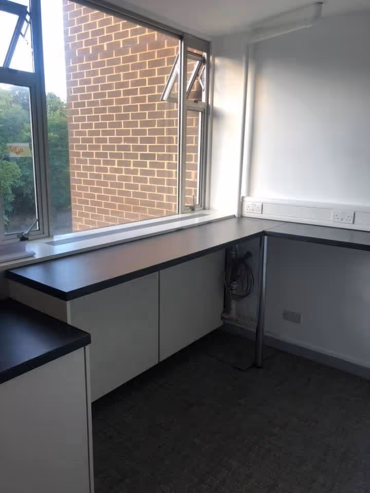 Empty corner office space with black countertops, white cabinets, two open windows, and multiple electrical outlets on the walls.