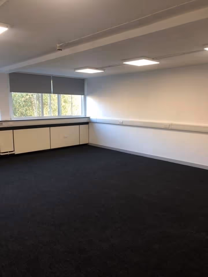 Empty office room with dark carpet, white walls, ceiling lights, and a window with closed blinds.