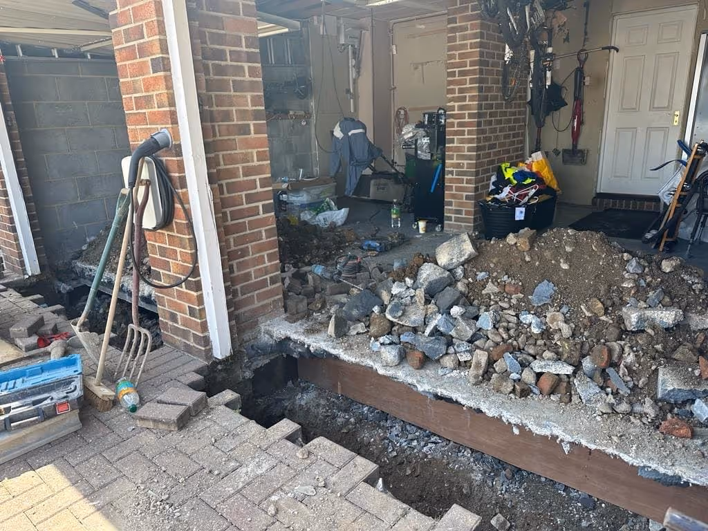 Construction site with excavated ground and rubble inside a residential garage area with tools and household items.