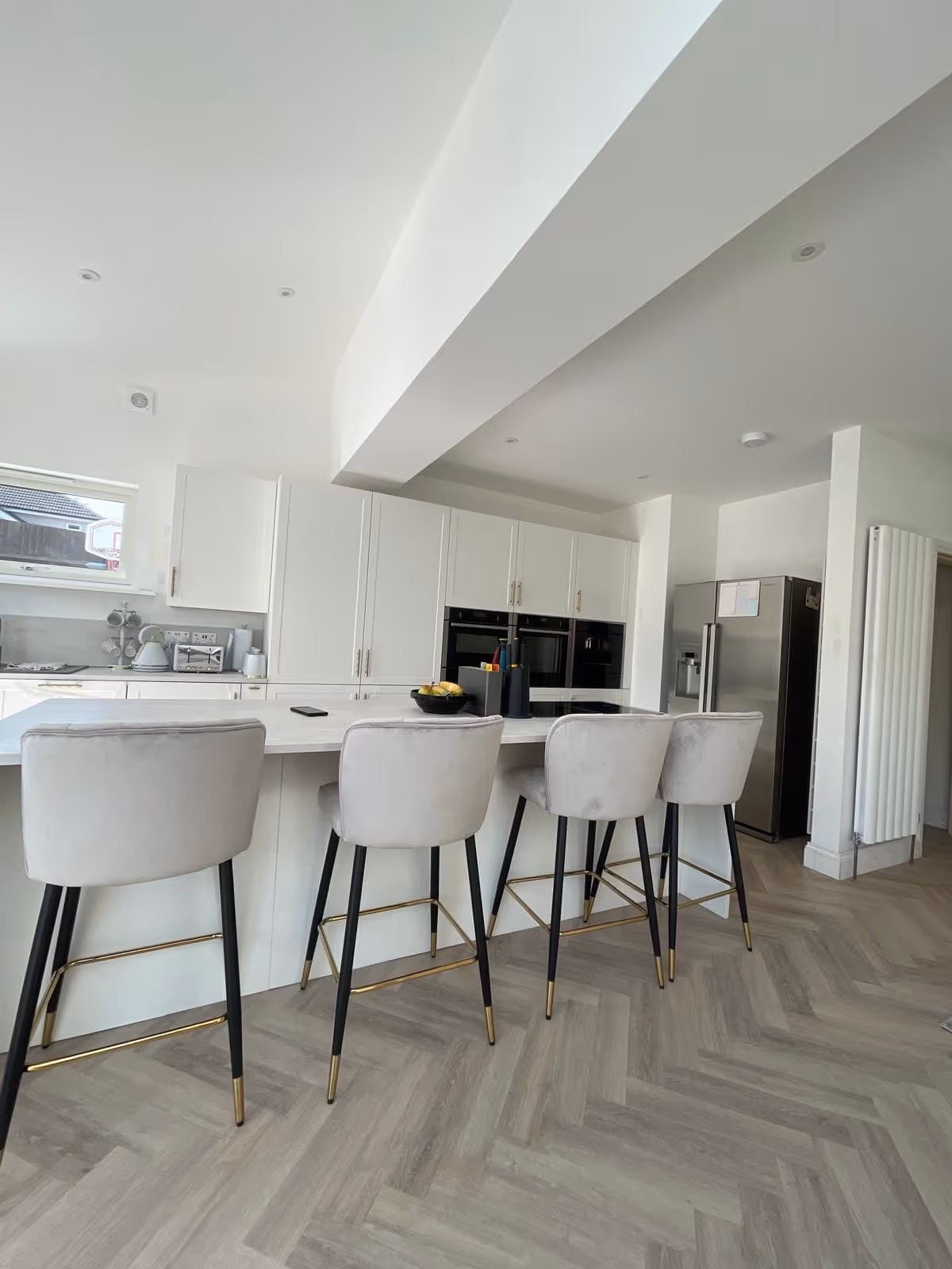 Modern kitchen with white cabinetry, a marble island countertop, four grey bar stools with black and gold legs, and stainless steel refrigerator.