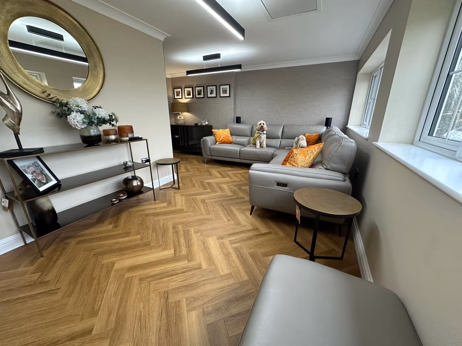 Modern living room with wooden herringbone floor, gray sectional sofa with orange pillows, two small dogs on the sofa, and a console table with decorative items and a round mirror.