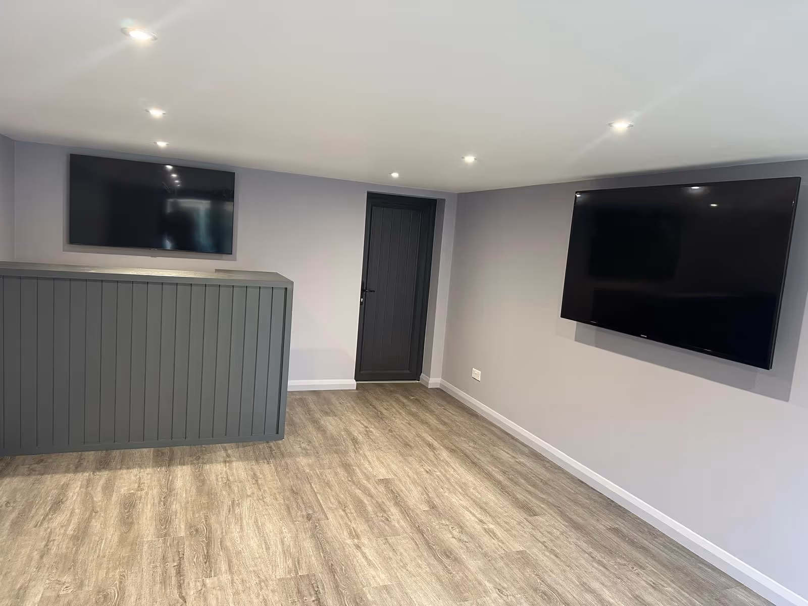 Empty modern room with light gray walls, wood flooring, two wall-mounted flat-screen TVs, a dark gray door, and a gray cabinet.