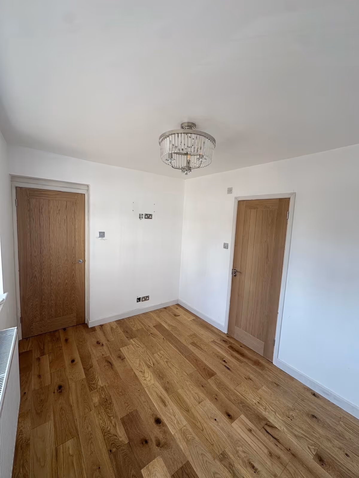 Empty room with light wooden flooring, two wooden doors, white walls, and a modern ceiling light fixture.