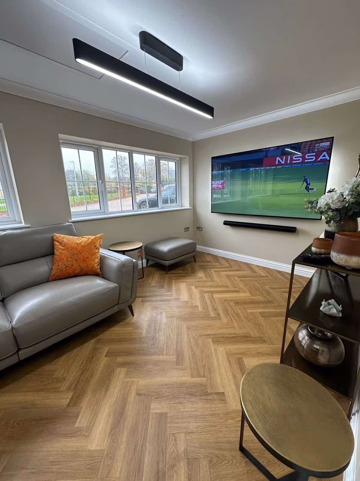 Modern living room with gray leather sofa, orange pillow, wall-mounted TV showing a cricket match, wooden herringbone floor, and contemporary furniture.