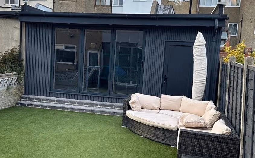 Modern black garden room with large windows and door, artificial grass lawn, and wicker outdoor sofa with cream cushions and a covered umbrella beside it.