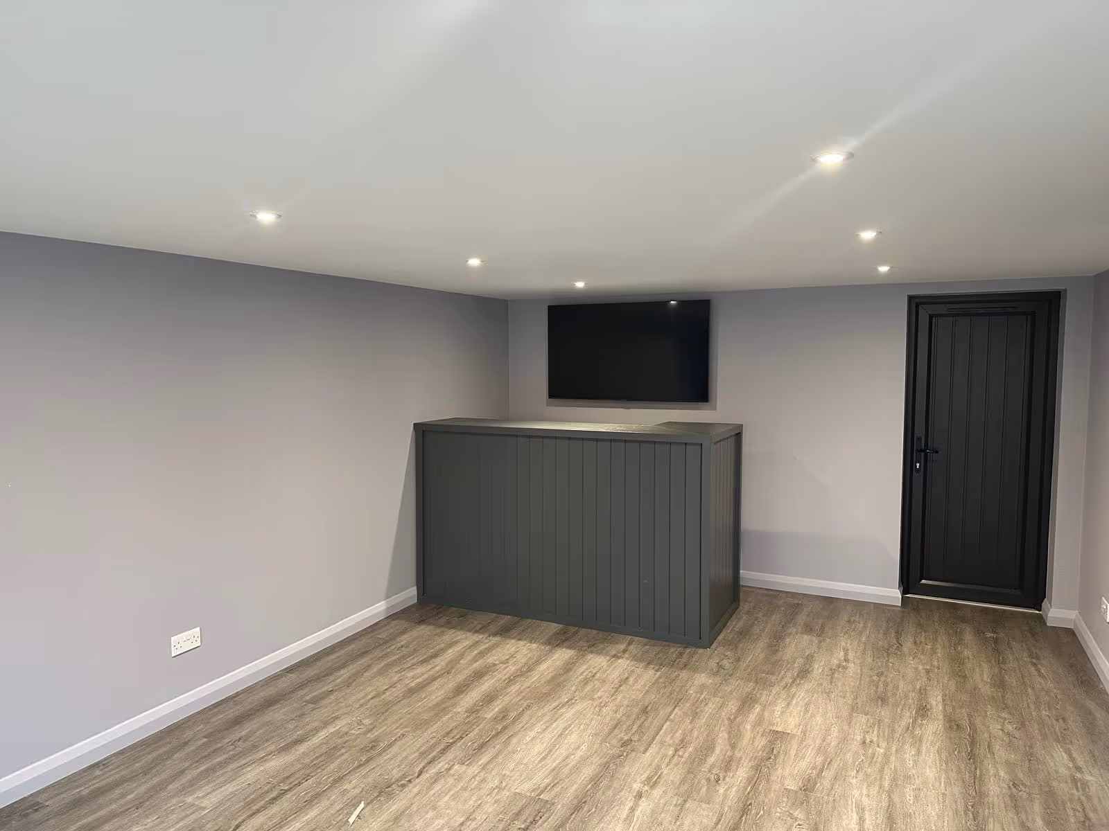 Minimalist room with light gray walls, wooden floor, a black reception desk, wall-mounted TV, and a black door.