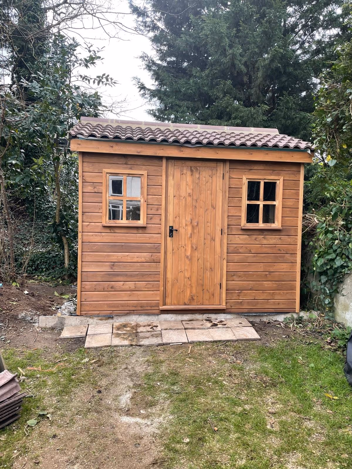 Wooden garden shed with a tiled roof and two small windows on either side of a central door, surrounded by trees and greenery.