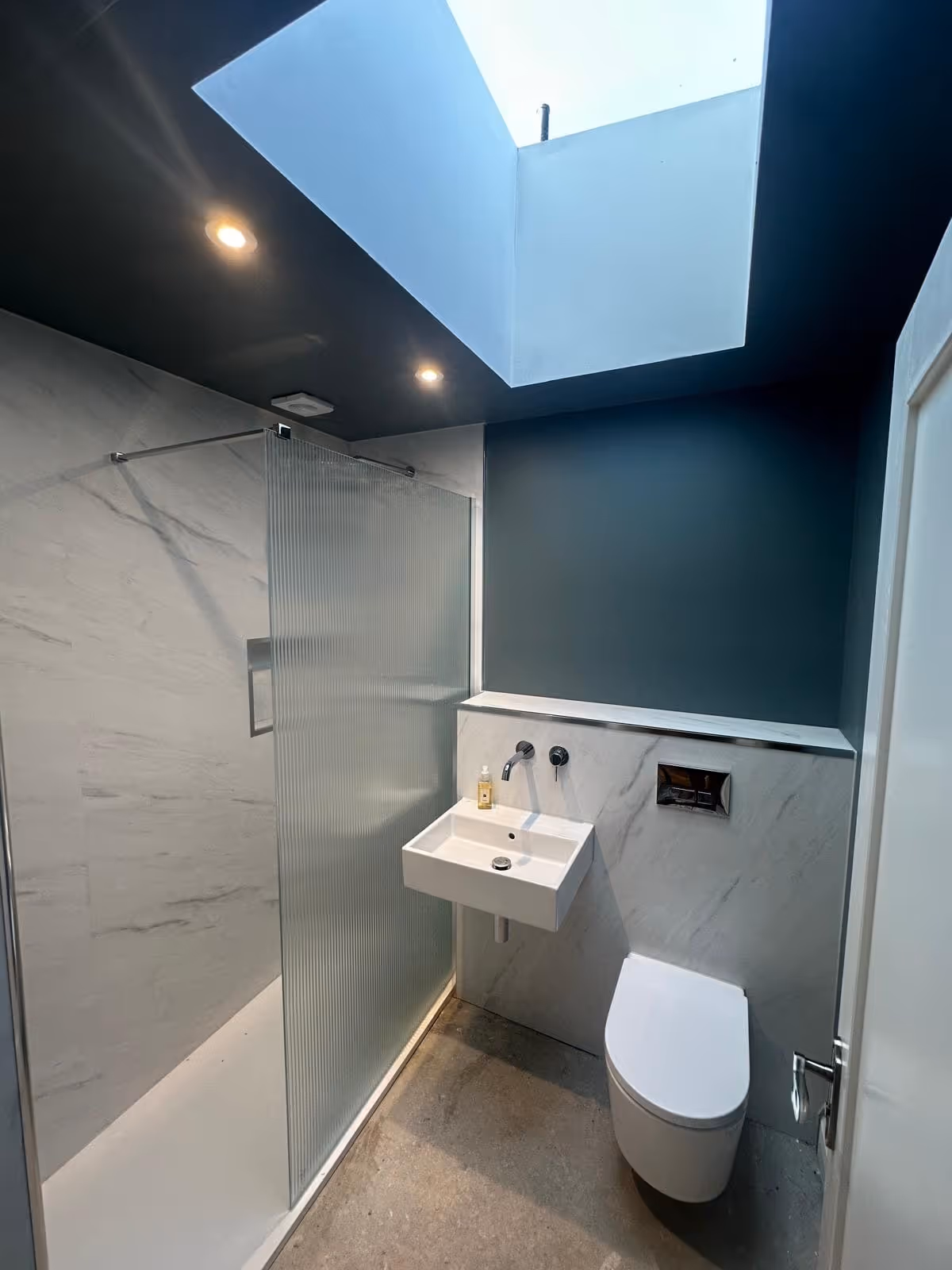 Modern bathroom with a skylight, walk-in shower with ribbed glass panel, wall-mounted sink, and wall-mounted toilet.