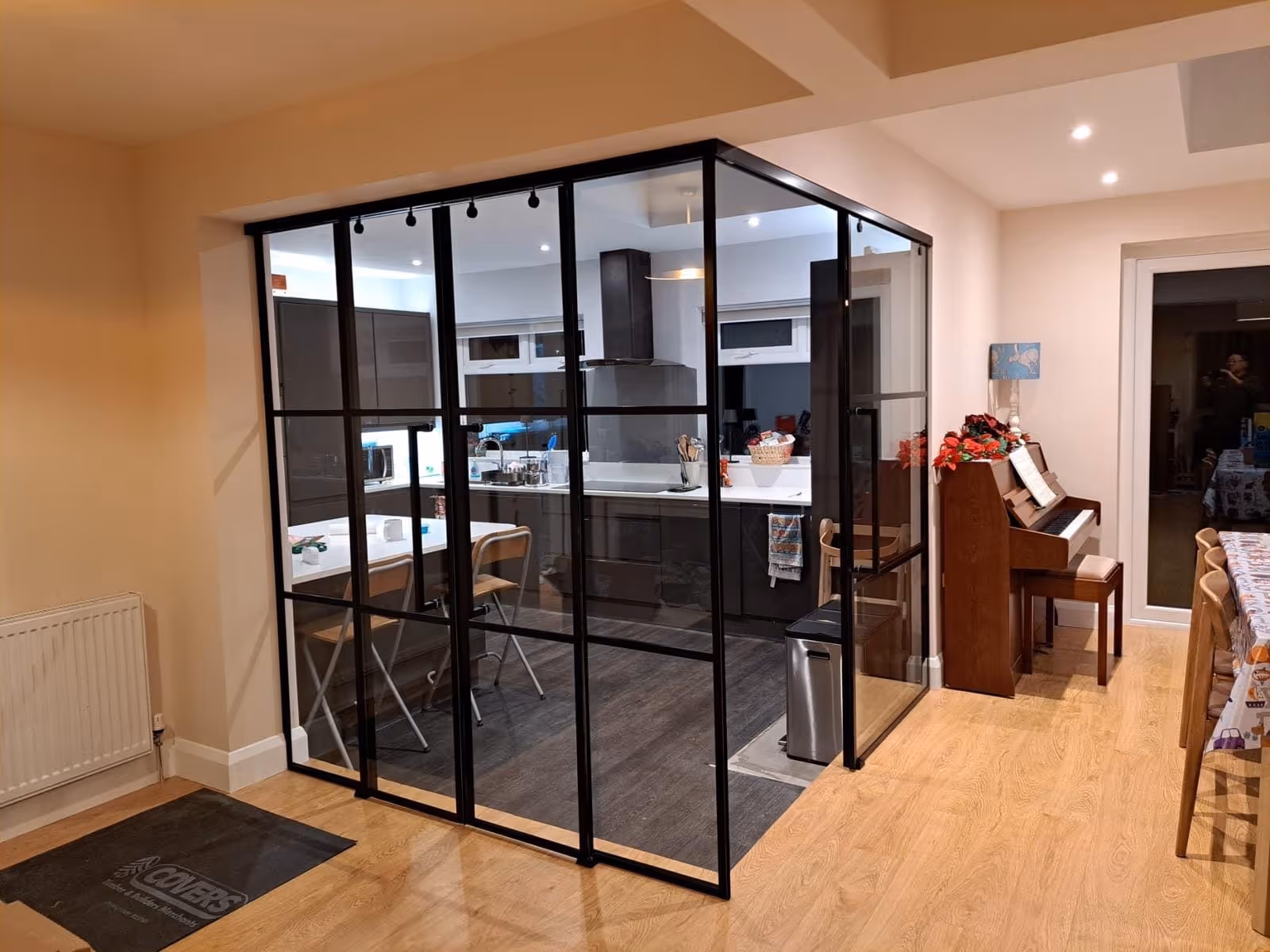 Modern kitchen enclosed by black-framed glass walls with wooden floor, adjacent to a dining area with a piano and floral lamp.