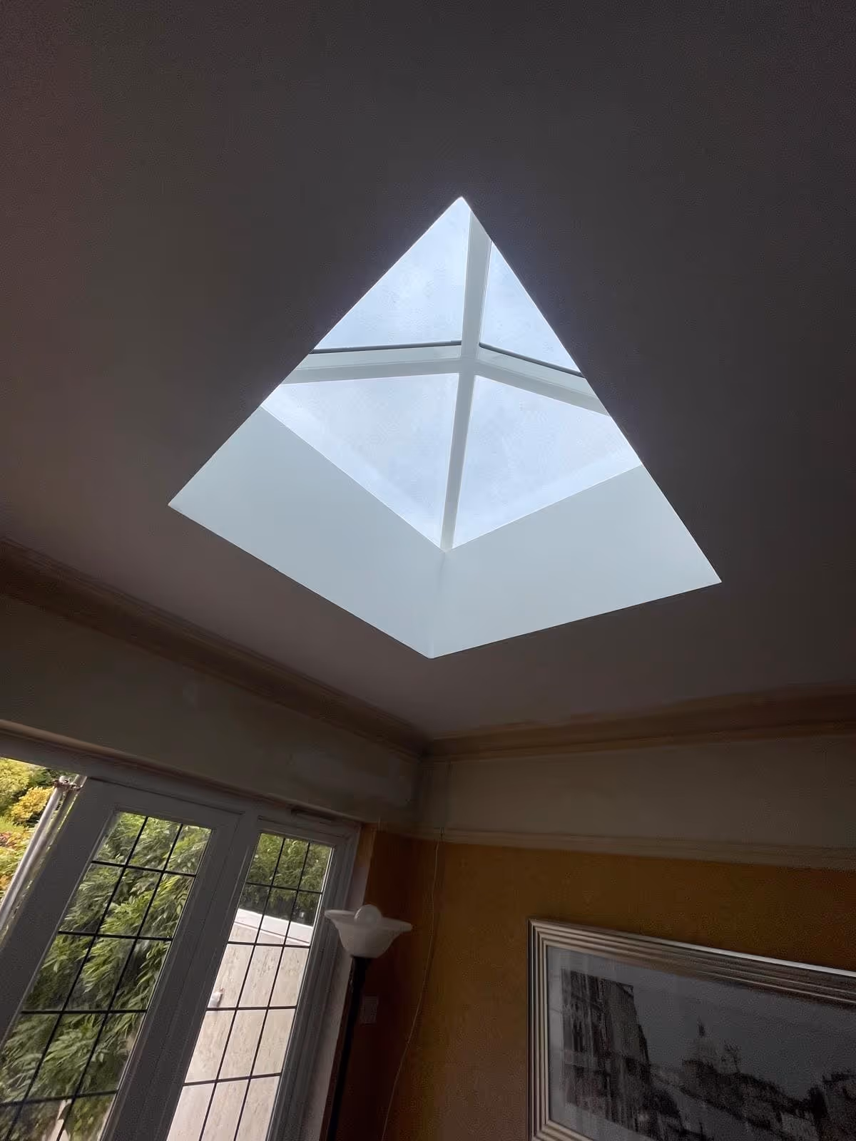 Interior view of a square skylight with four glass panes on a ceiling, above a room with a window and framed artwork.