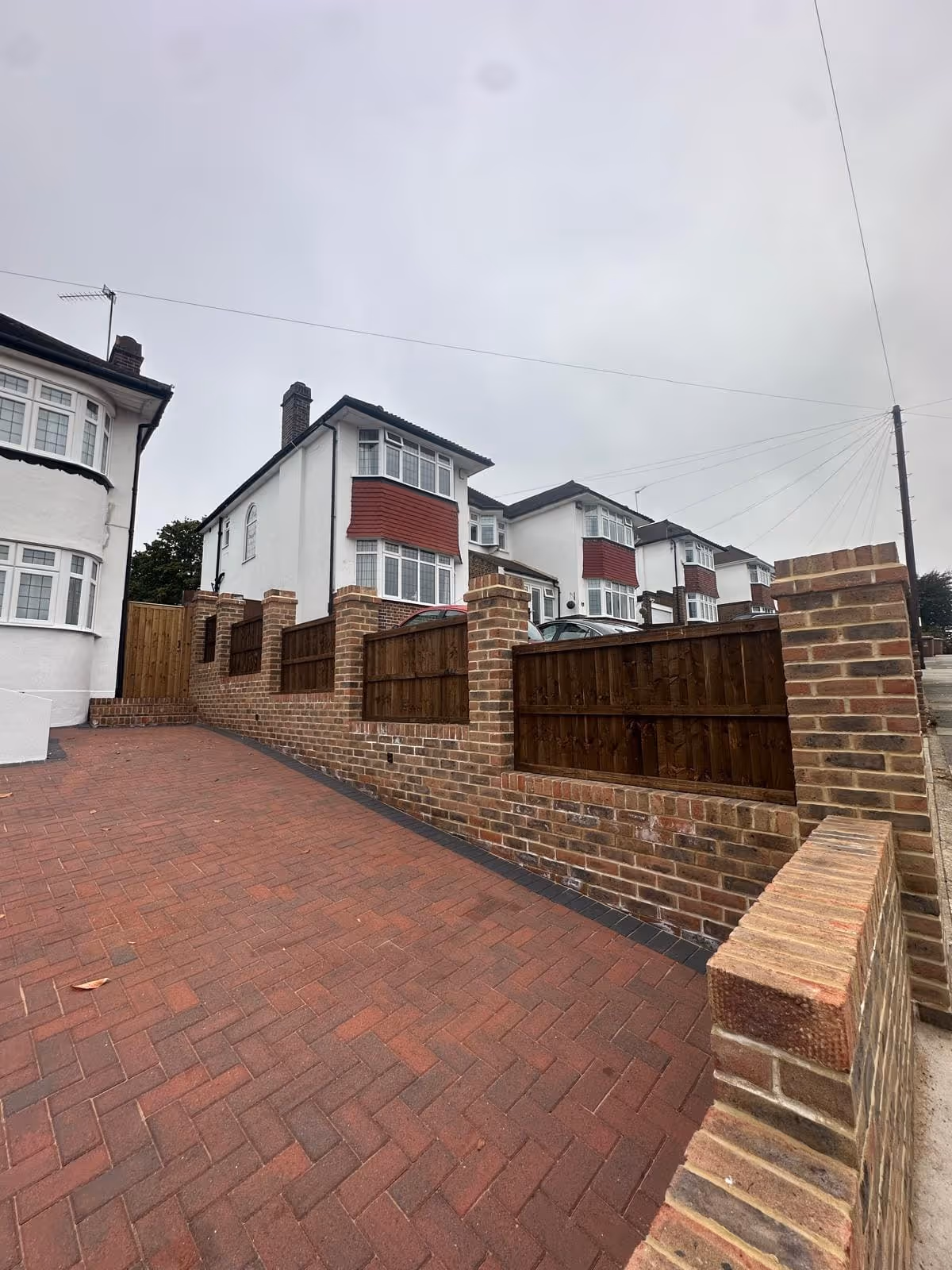 Driveway with red brick paving leading to white houses with red bay windows and brown wooden fence panels on brick pillars.
