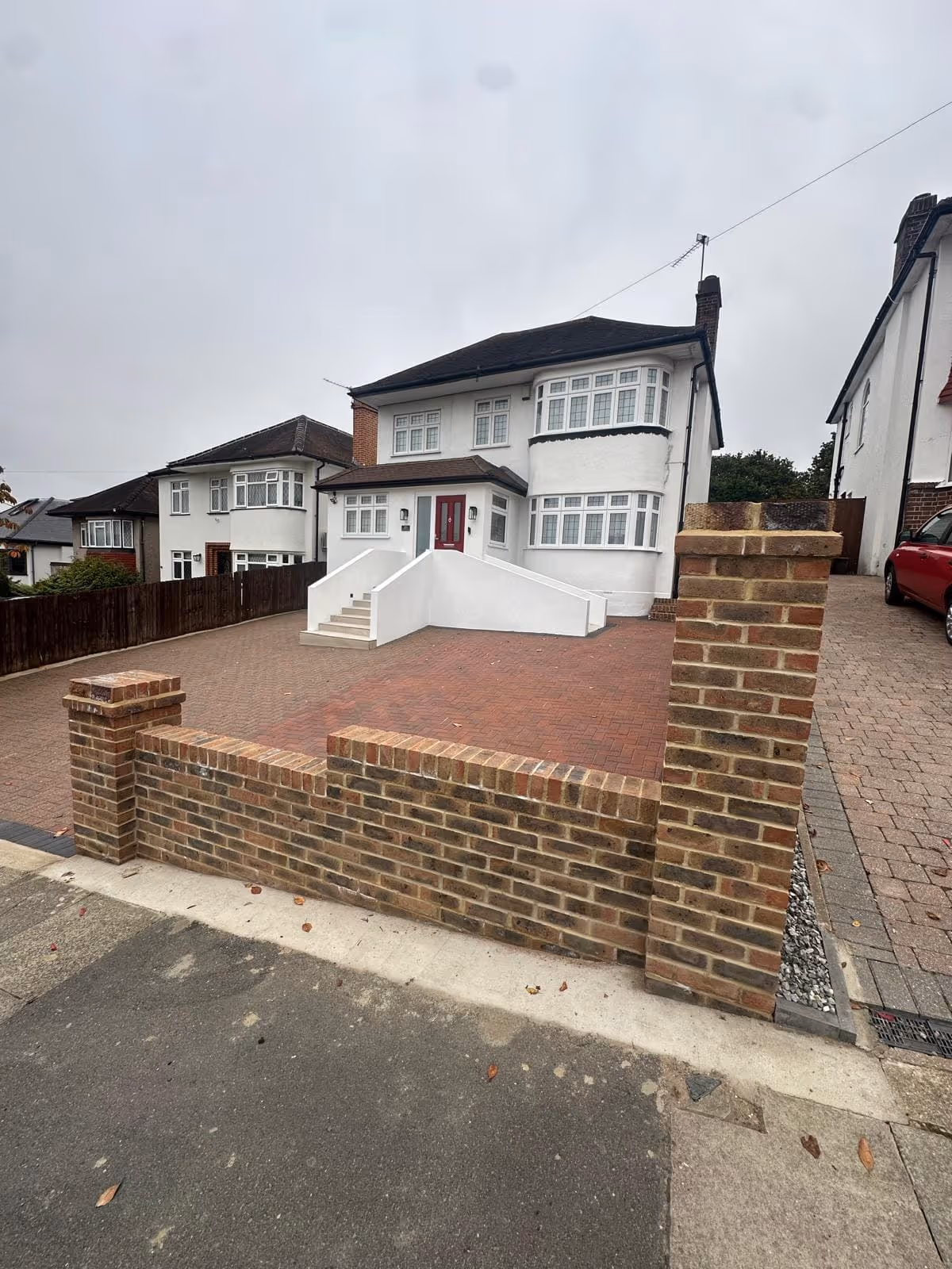 Two-story white house with a red front door, brick driveway, and a brick fence with two pillars under a cloudy sky.