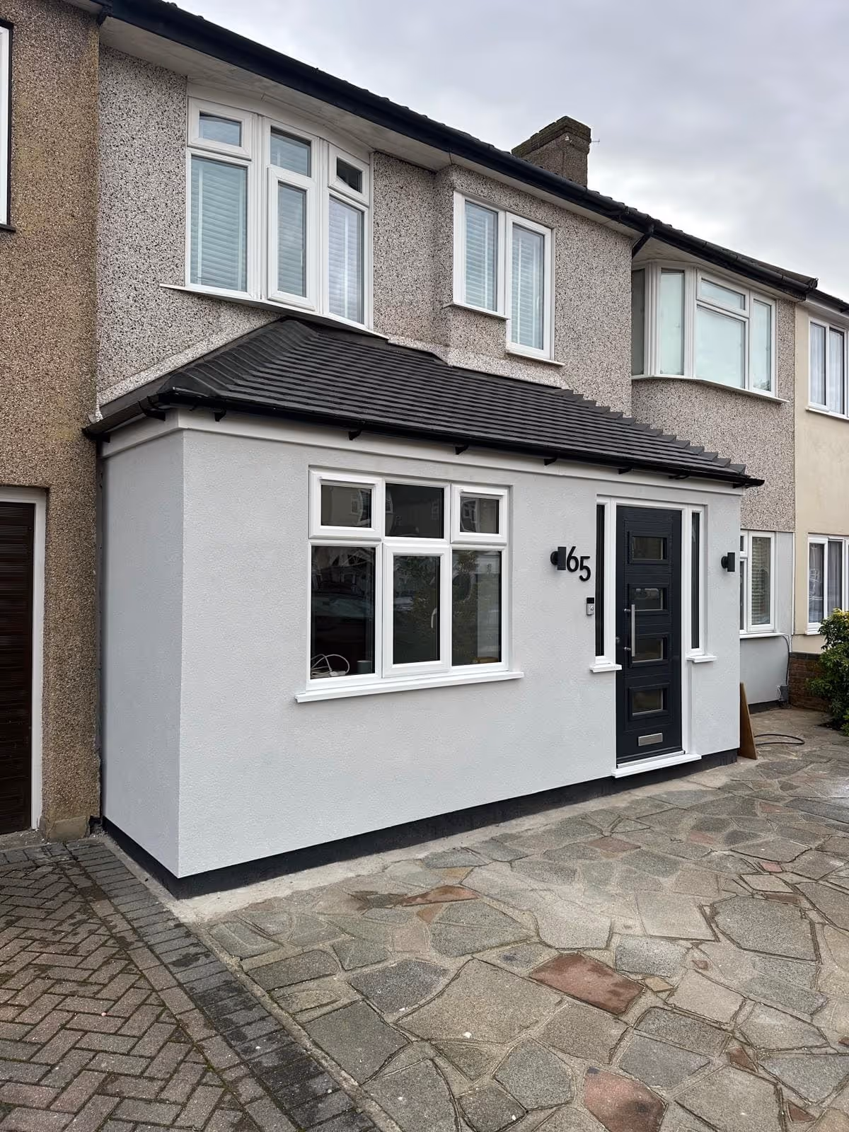 Two-story house with a gray stone driveway, light gray extension, black door, and white-framed windows under an overcast sky.