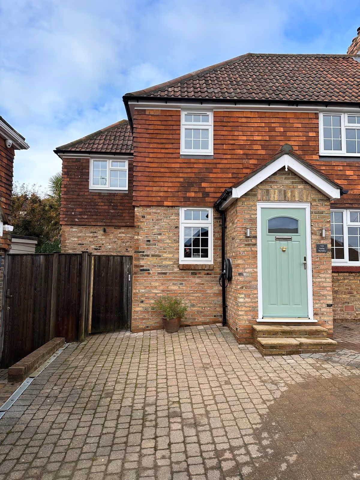 Brick and tile house with a light green front door, white-framed windows, a paved driveway, and a wooden gate on the left.