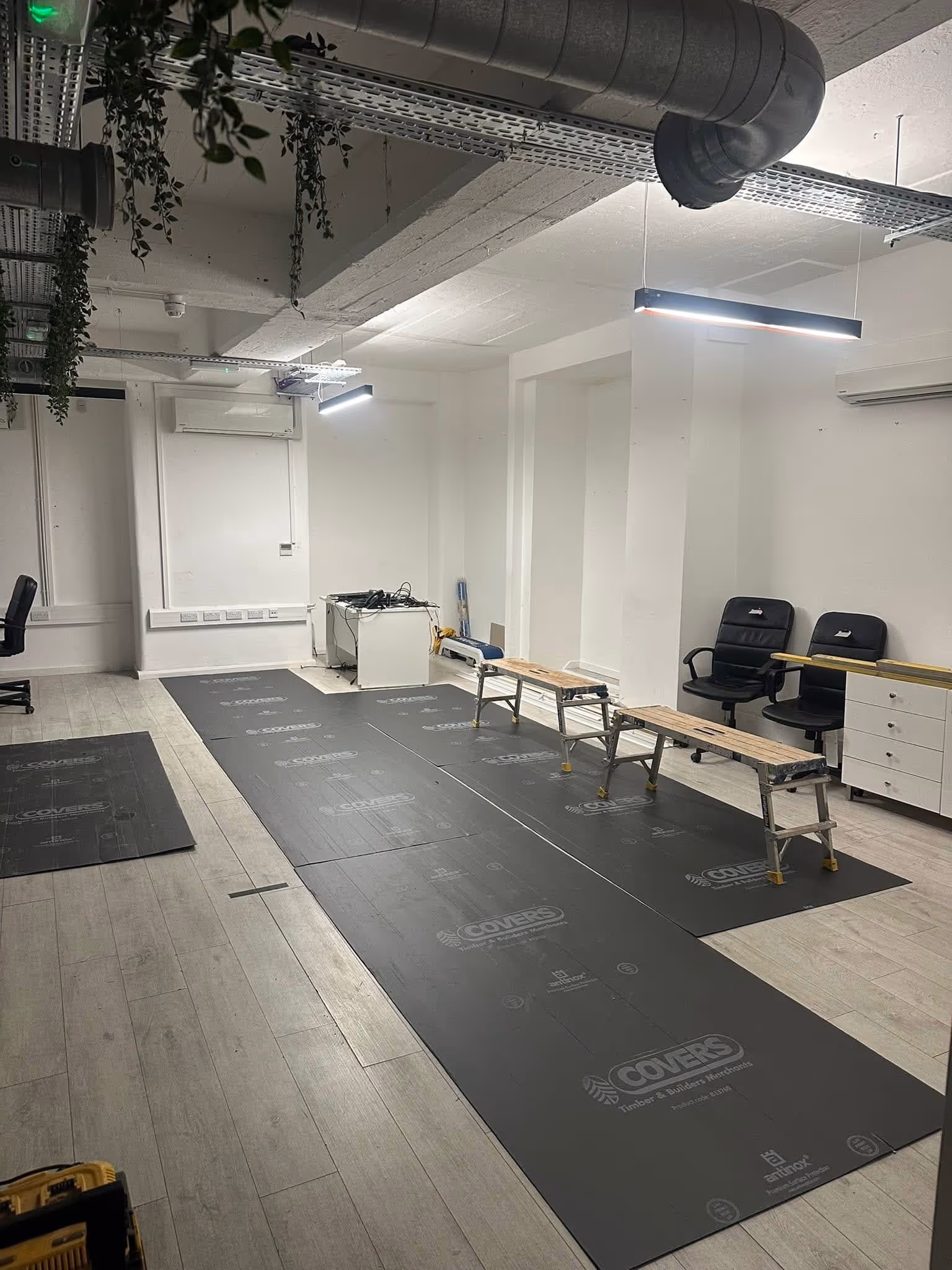 Office space under renovation with black protective floor sheets, two wooden benches, and two black office chairs.