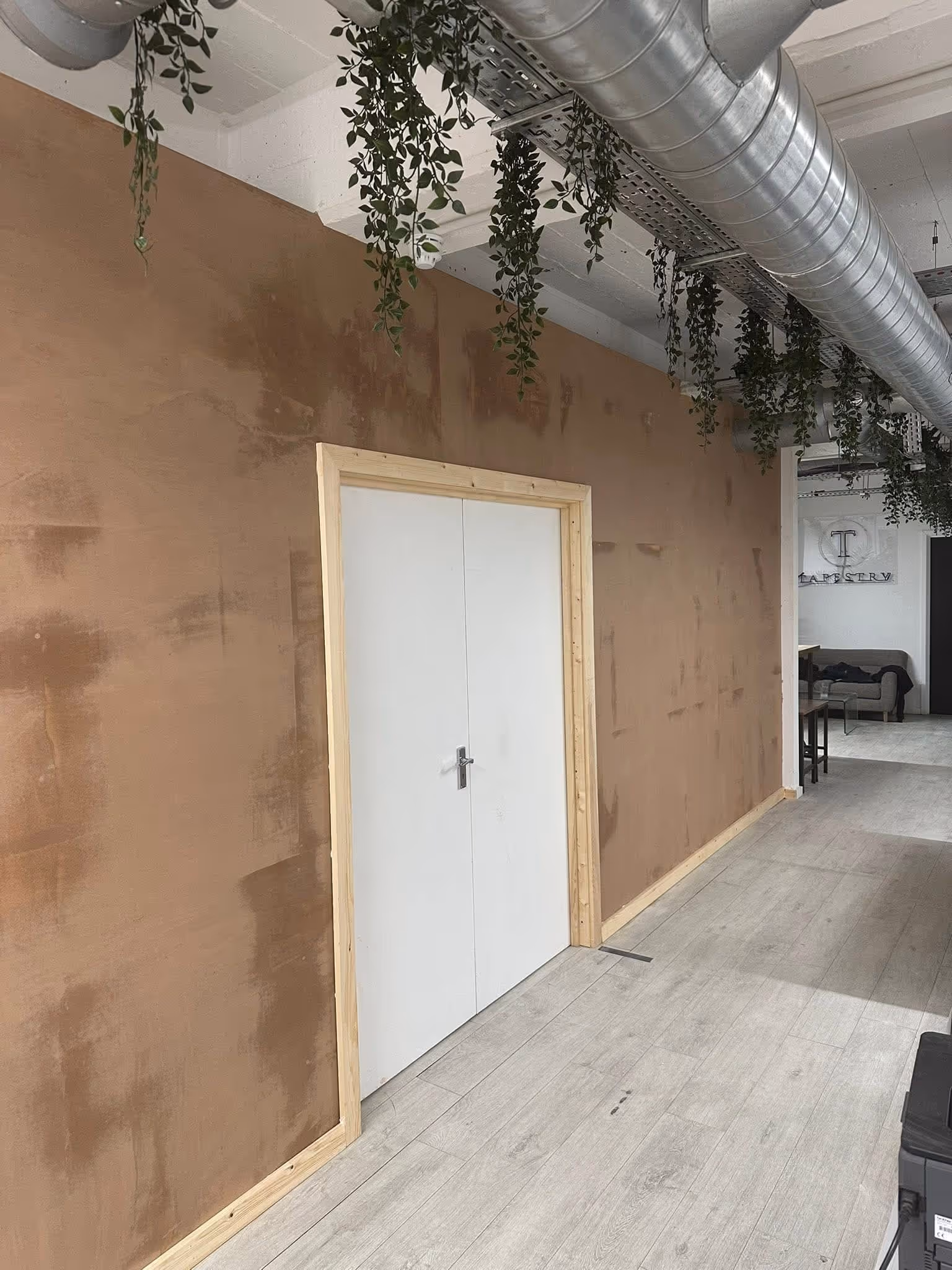 Interior hallway with a double white door framed by natural wood, brown textured wall, hanging green plants, and visible silver air ducts on the ceiling.